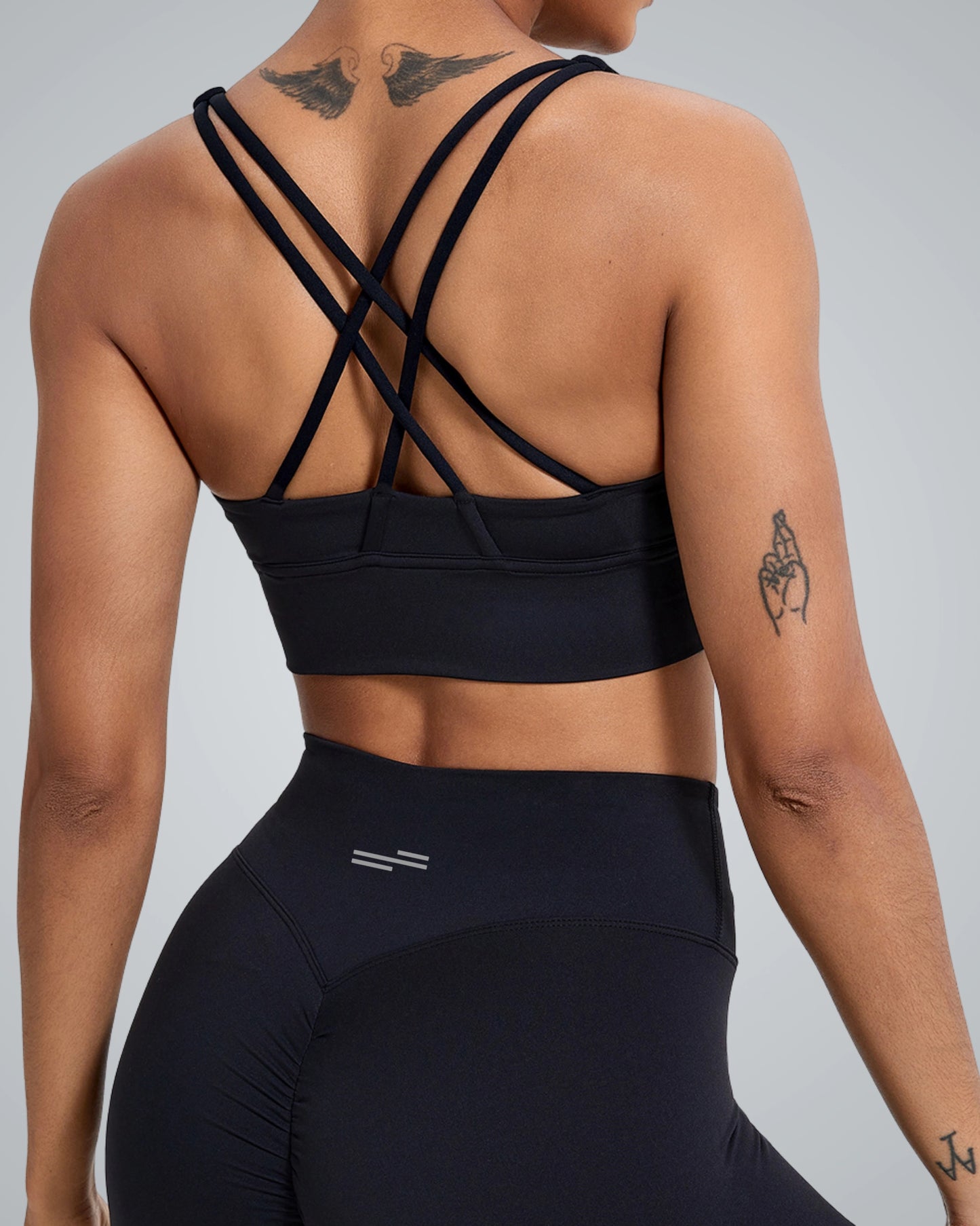 A woman wearing a Bandsam black sports bra with crisscross straps at the back, showcasing her shoulder tattoos. The image focuses on her torso and back, highlighting the activewear's design and fit.