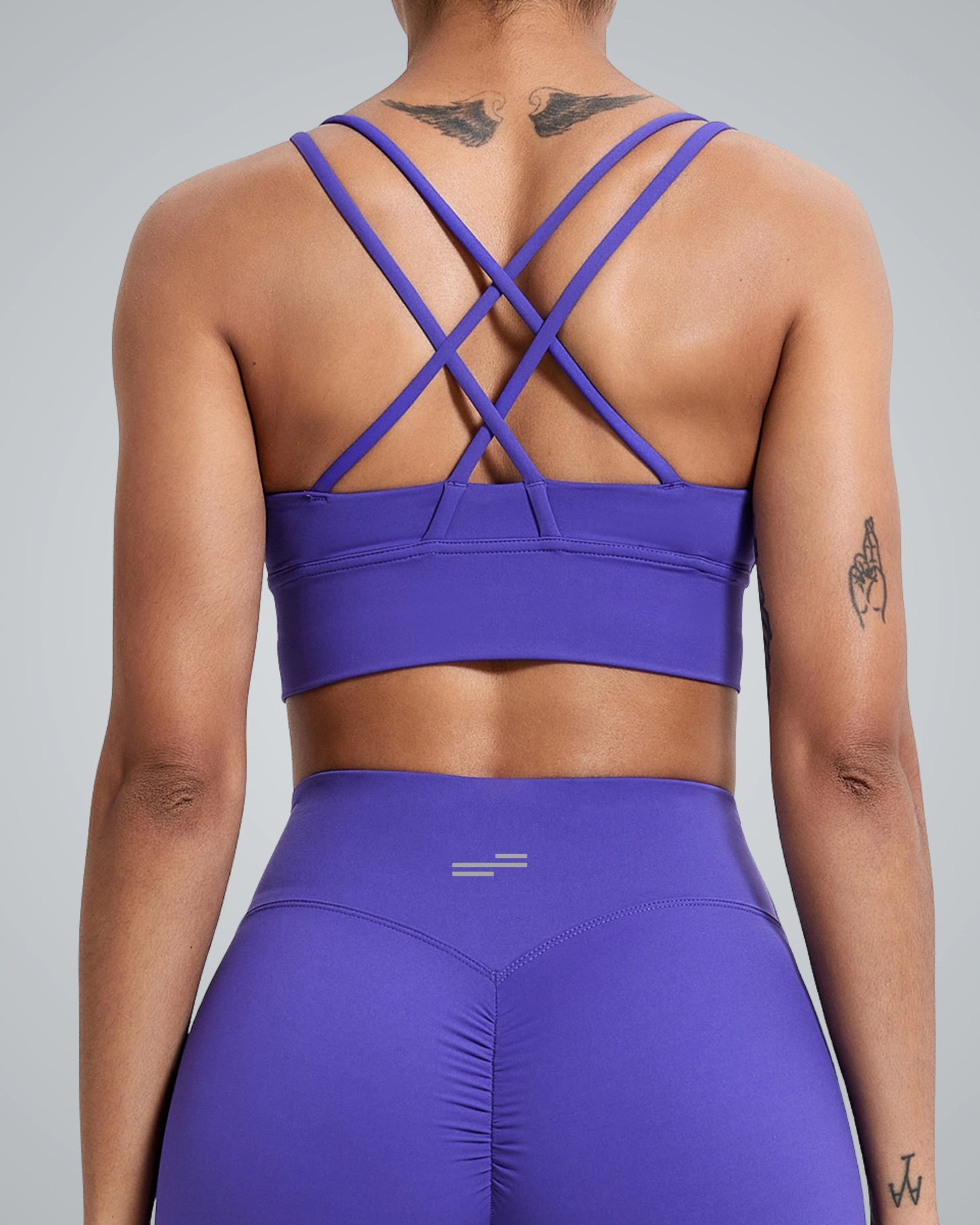 A women wearing a purple breathable Bandsam workout bra with a crisscross design and matching high-waisted leggings, shown from the back. Tattoos are visible on their upper back, arm, and wrist.