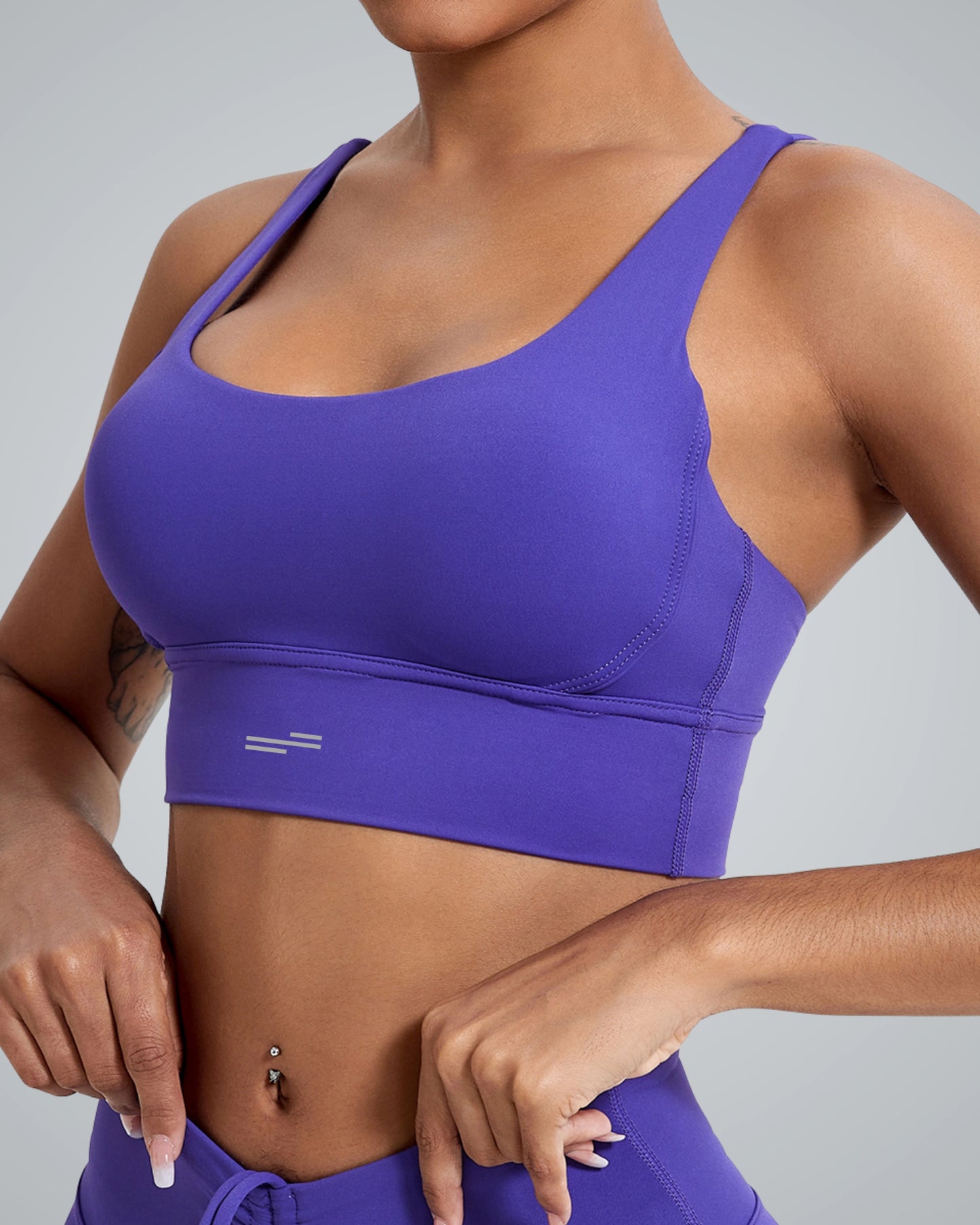 A women wearing a Bandsam purple FlexFit Bra and matching bottoms stands against a gray background, showing off the bras stylish crisscross design, a tattoo on their left arm, and a belly button piercing.