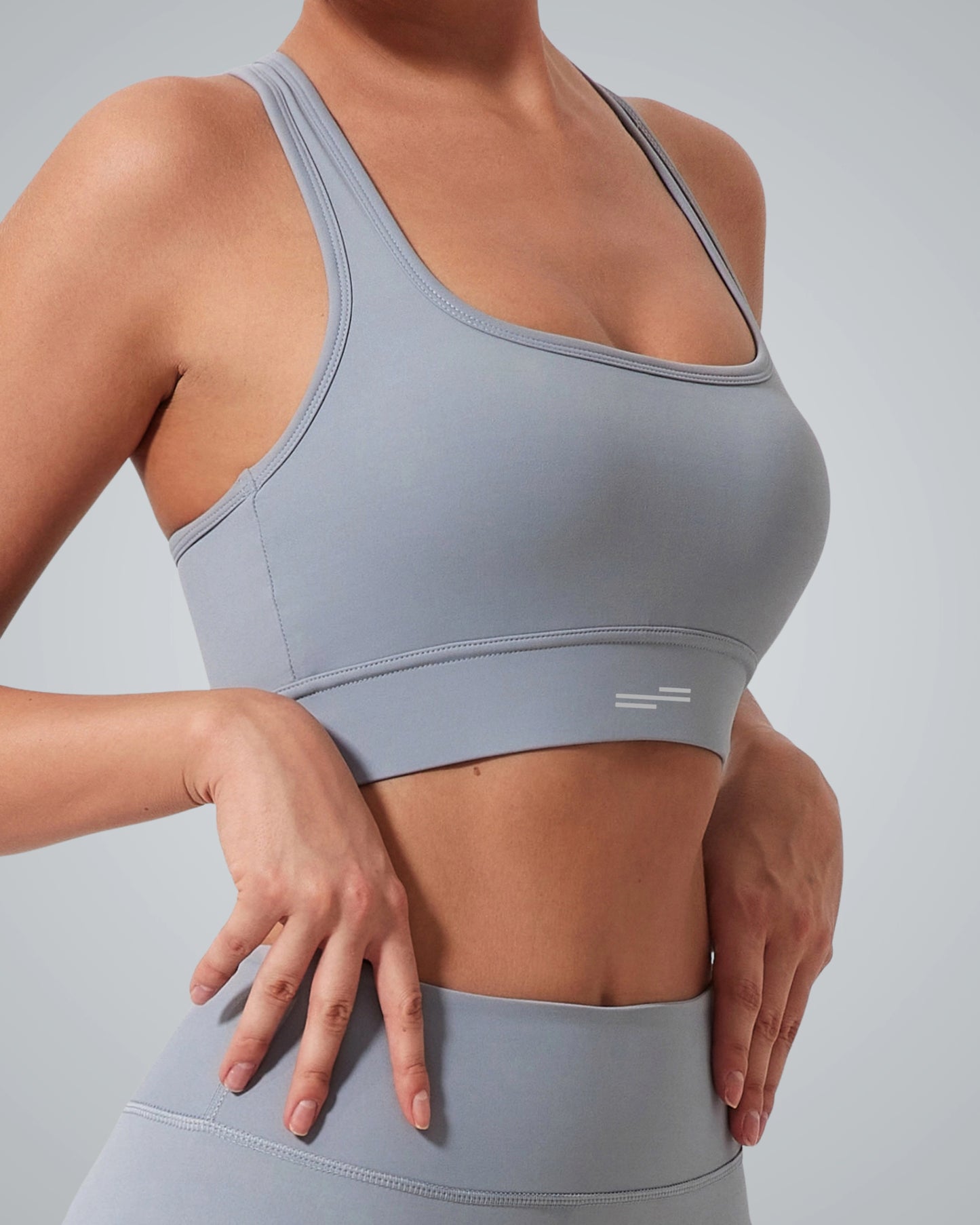Woman wearing a Bandsam GlowMotion Bra, a high impact sports bra with exceptional support, and matching high-waisted leggings, posing with hands near her waist against a plain light gray background.