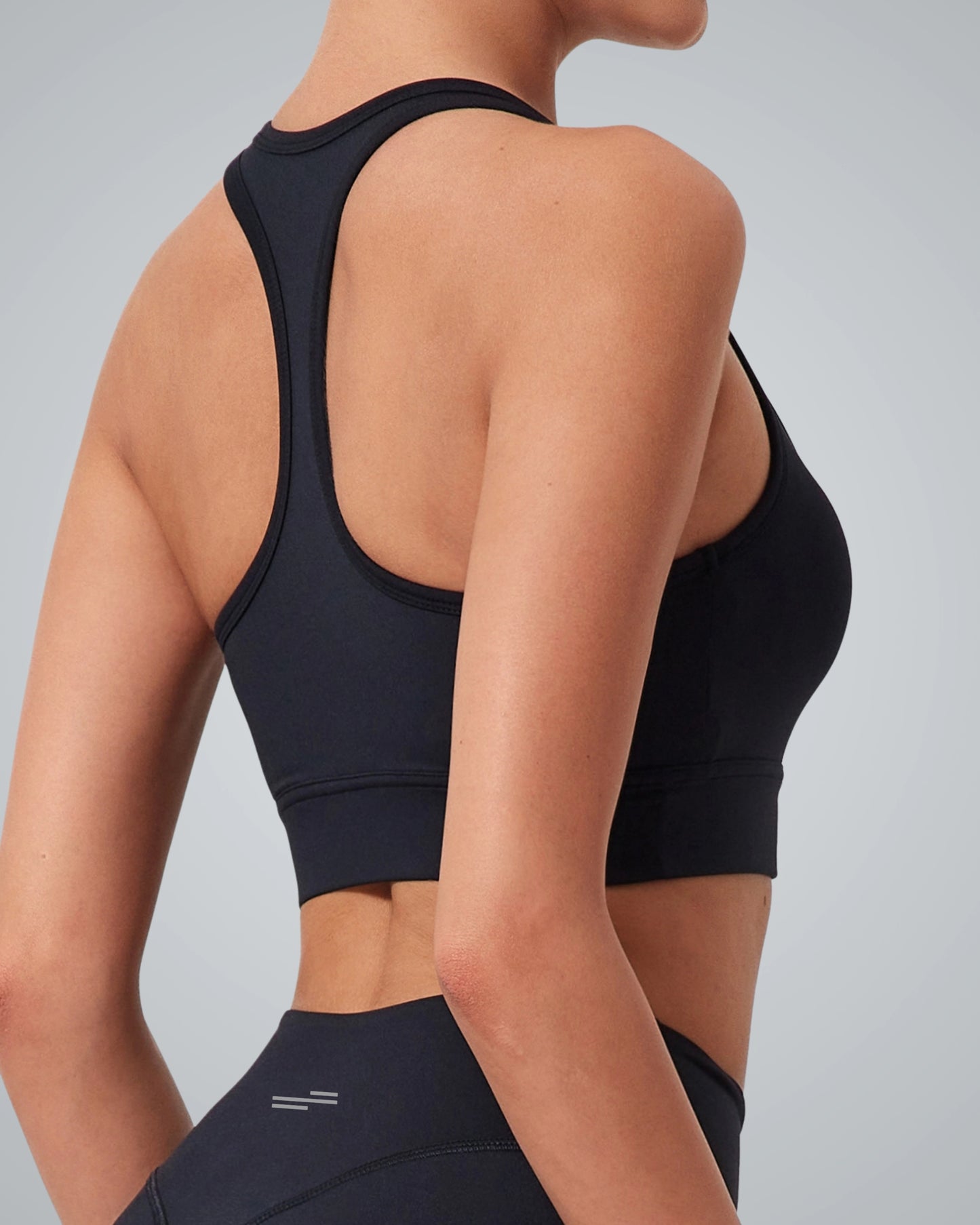 The Bandsam GlowMotion sports bra in black, worn by a woman paired with high-rise leggings, back view highlights fit and design on an uncluttered gray background.