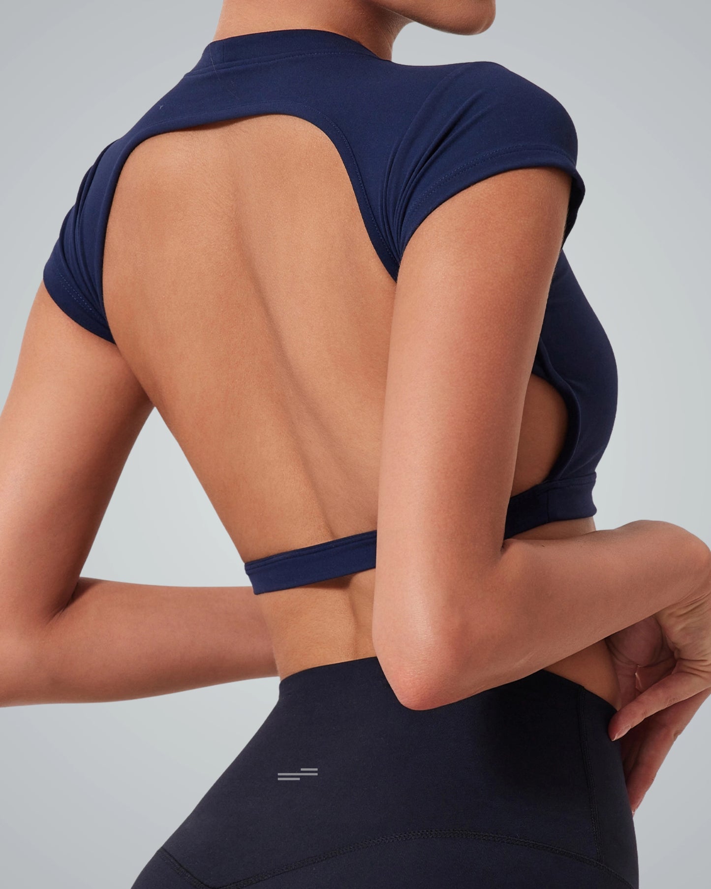 A woman wears a Bandsam navy blue, short-sleeved ScarletFlow Top with an open back and high-waisted black leggings, shown from behind against a plain background—a stylish choice for any exercise wardrobe.