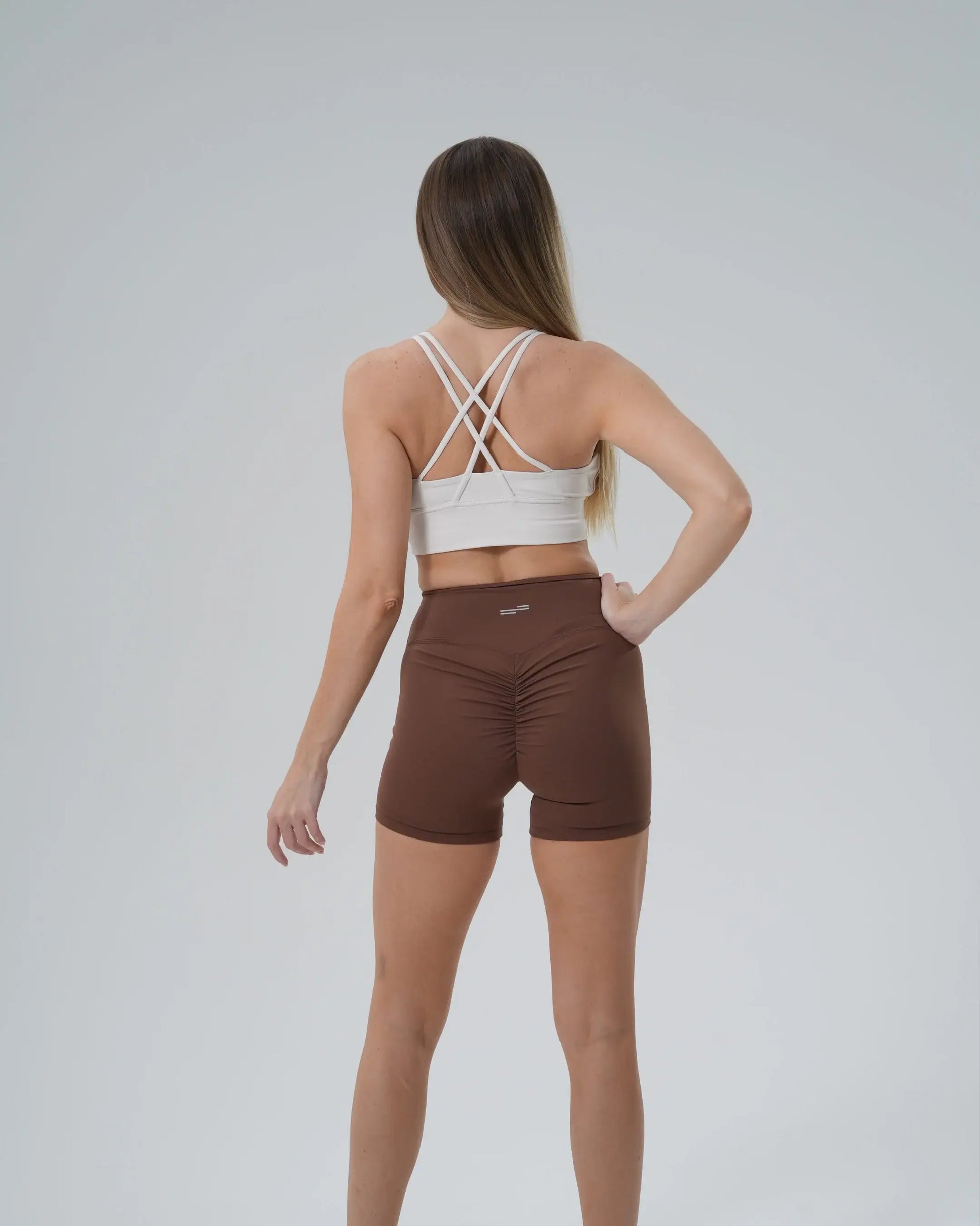 Back view of woman in Bandsam Edge Pro Set, featuring white sports bra and brown shorts, showcasing a minimalist and performance-focused design.