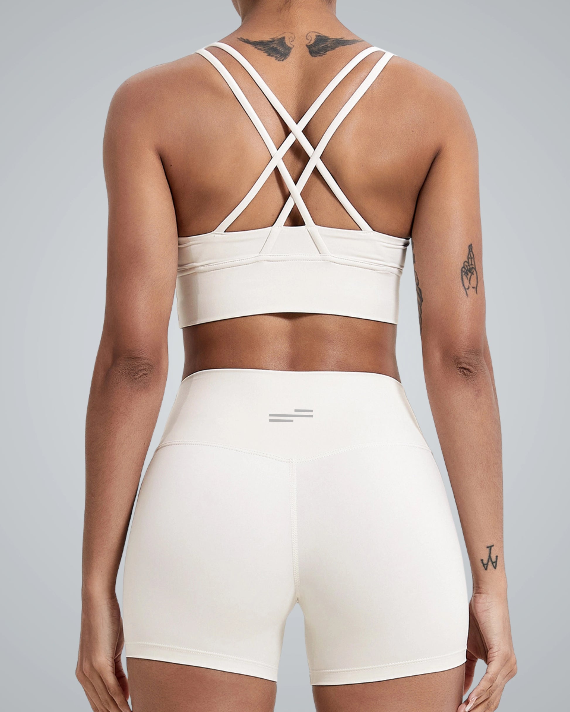 A women wearing a matching Bandsam white FlexFit Bra with a crisscross design and high-waisted shorts is shown from the back, displaying several tattoos on their back and arms against a plain gray background.