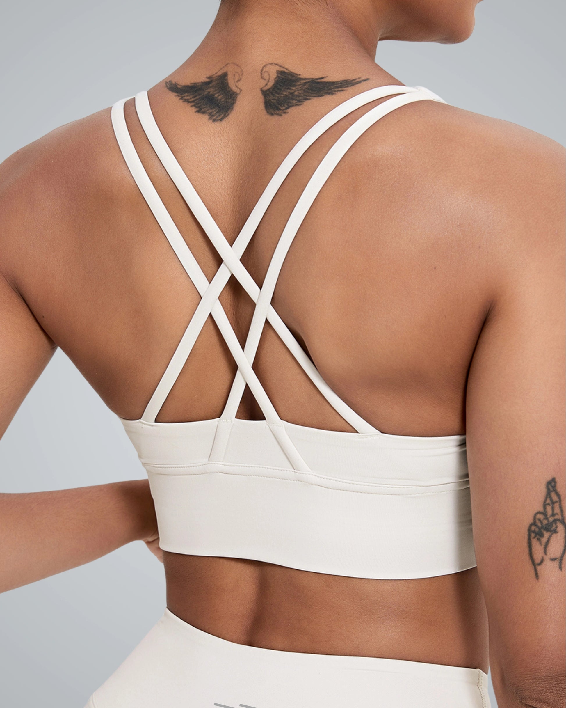 A women wearing a Bandsam white FlexFit Bra with a crisscross design on the back; visible wing and floral tattoos are on their upper back and left arm.