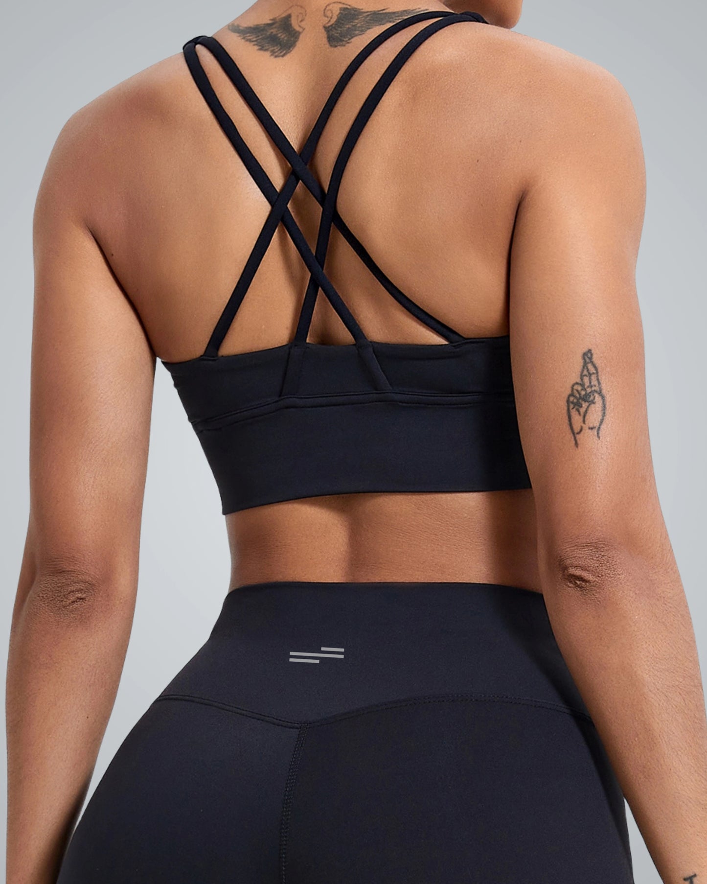 A woman wearing a Bandsam black sports bra with crisscross straps and matching high-waisted leggings, showing her back and shoulder tattoo against a plain gray background.