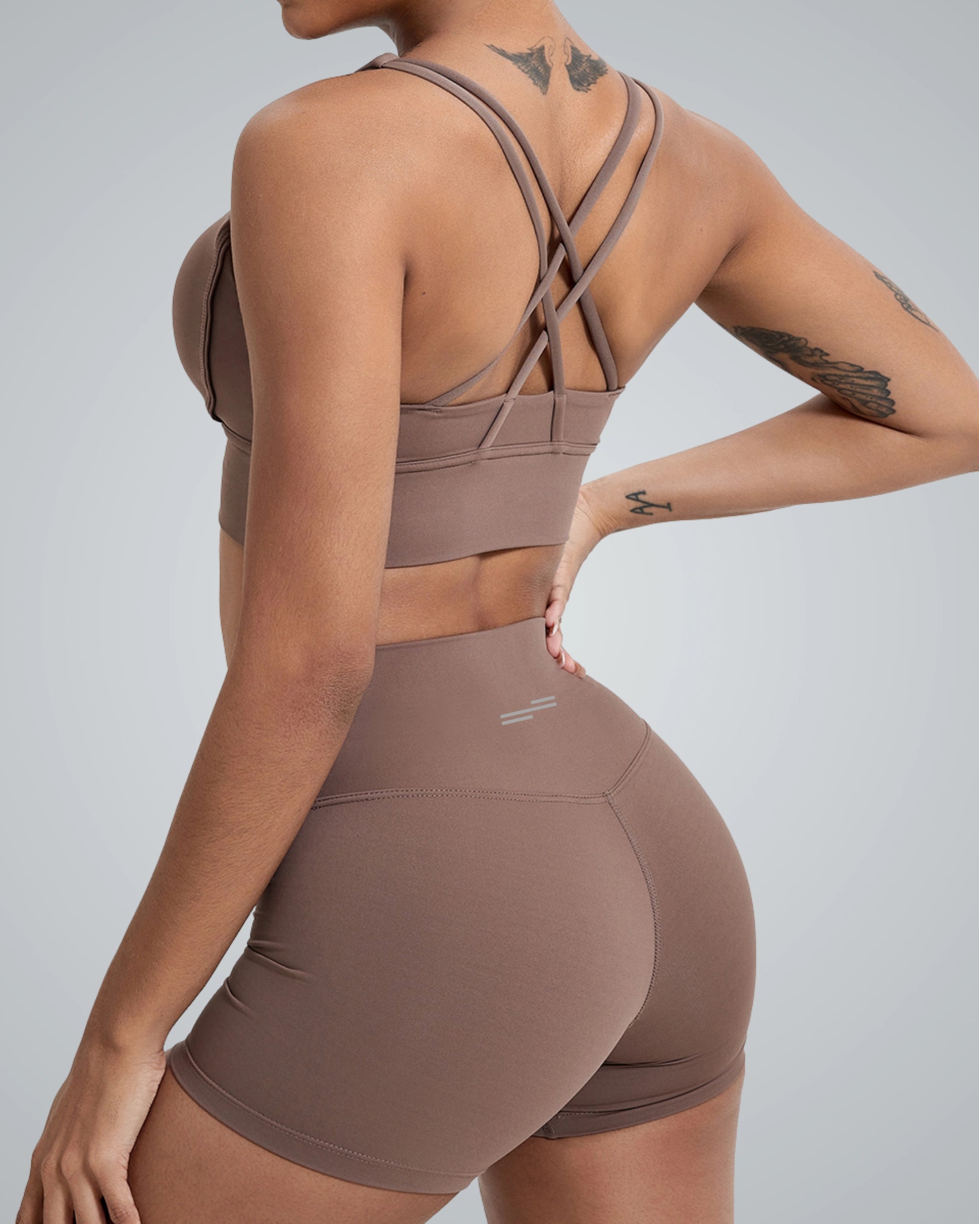 A woman wearing a Bandsam brown sports bra with crisscross straps and matching high-waisted shorts. She is facing away from the camera, showcasing a sleek and stylish athletic look against a neutral gray background.
