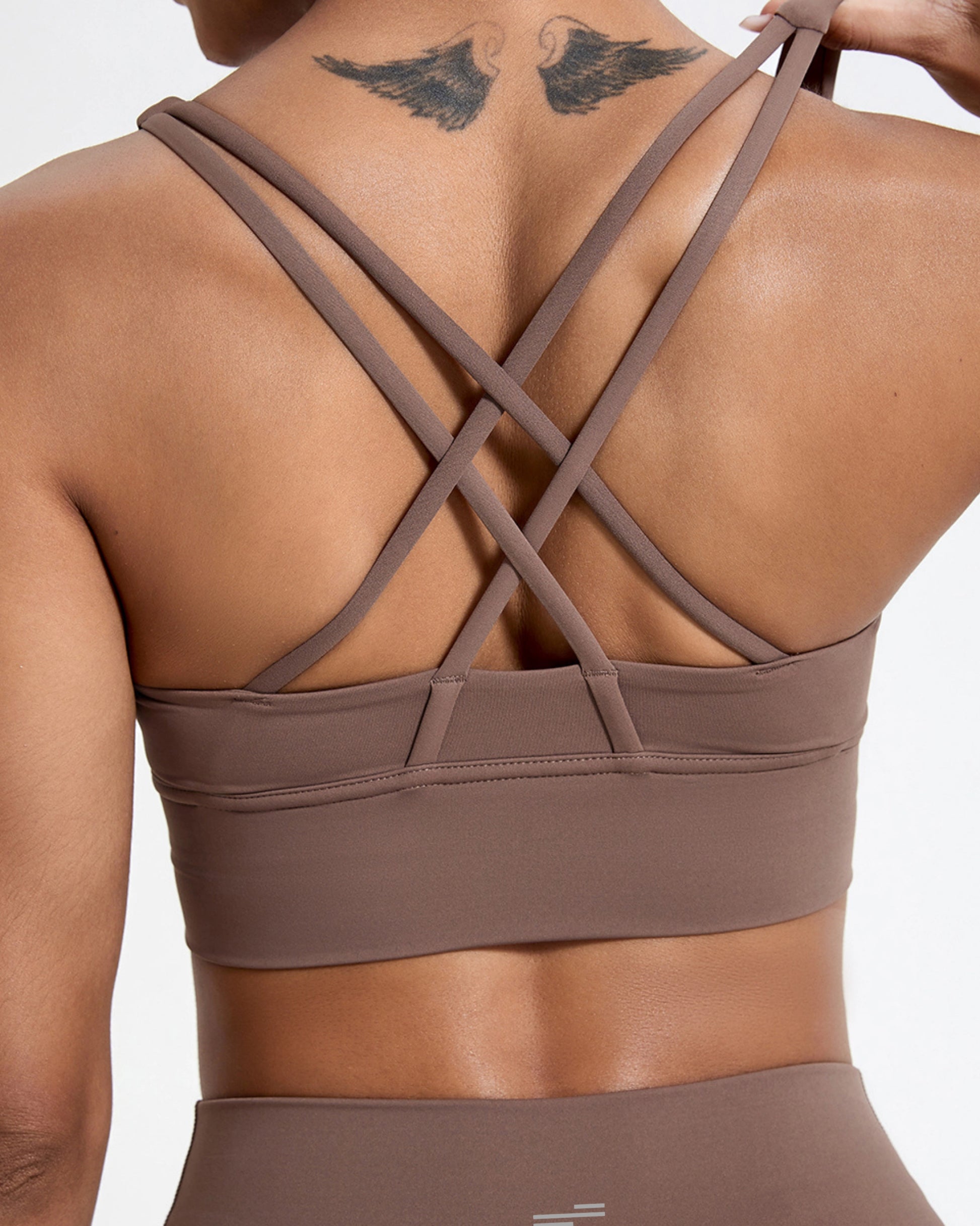 A close-up of a woman's back wearing a Bandsam brown sports bra with crisscross straps. The image also features a tattoo of bird wings on her shoulder, contrasting with her skin tone and the fabric of the bra.