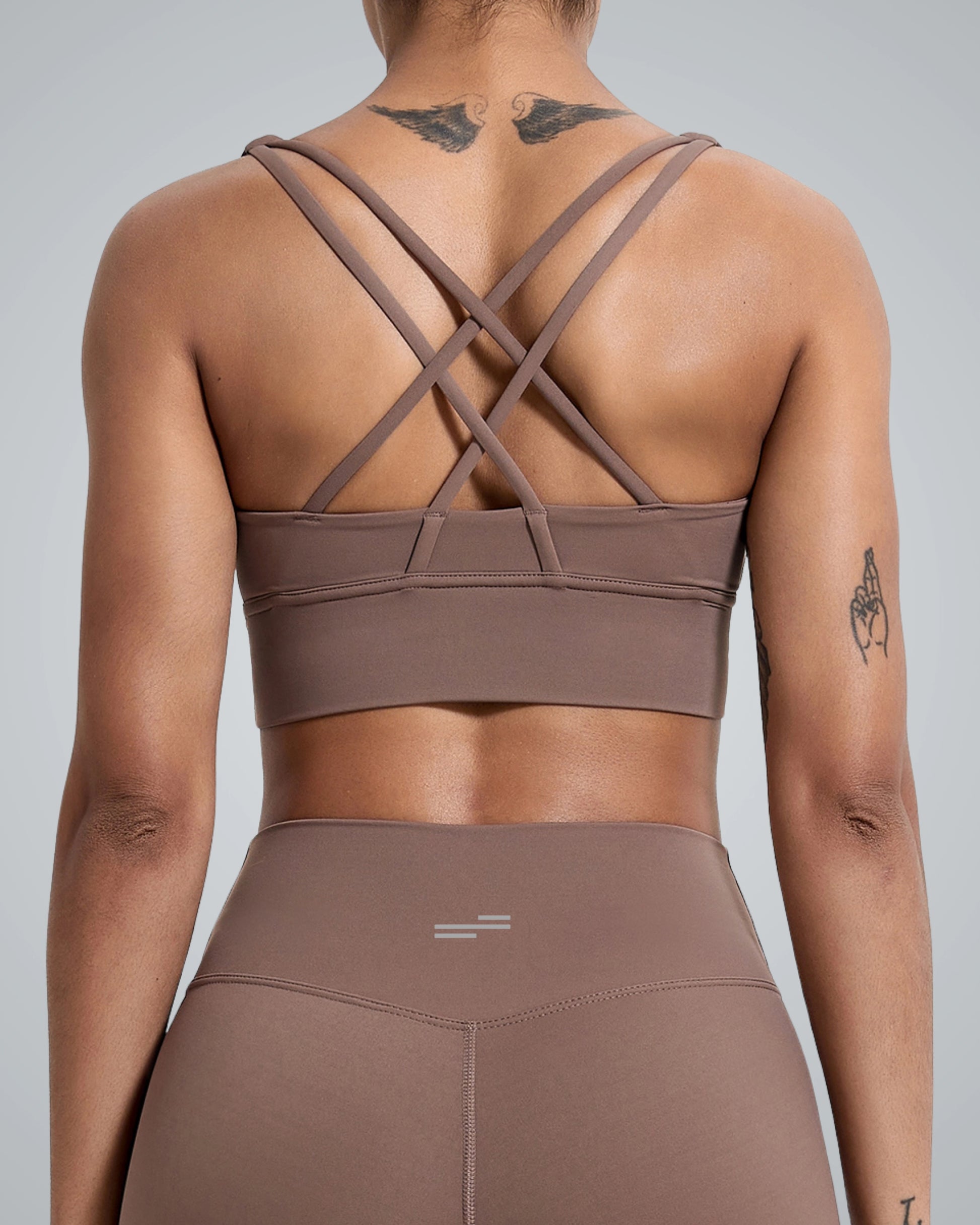 A close-up view of a women wearing a Bandsam brown sports bra with crisscross straps on the back, paired with matching high-waisted leggings. The person has visible tattoos on their upper back, and the background is a soft gray color.