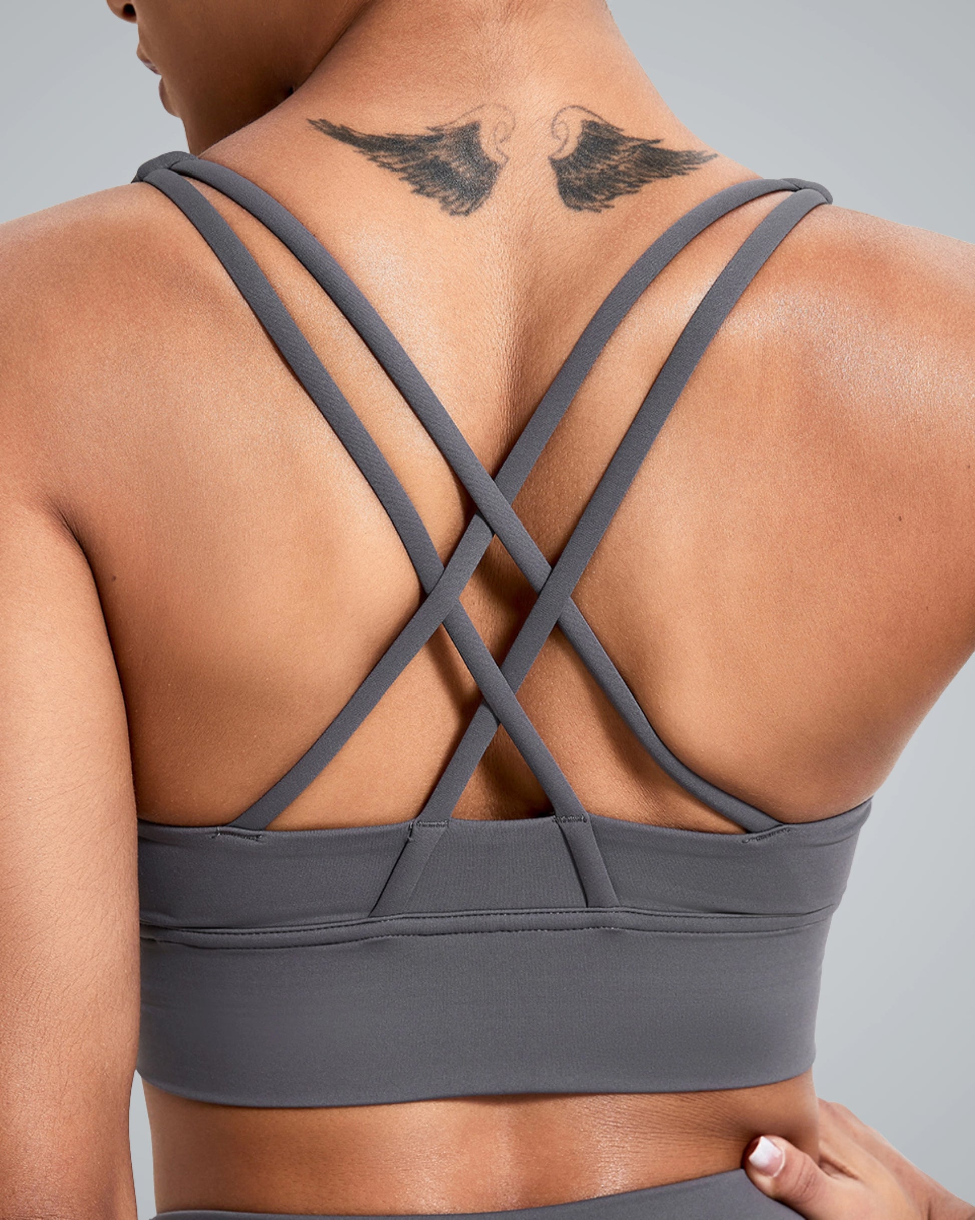 A close-up of a women wearing a Bandsam gray sports bra from the back, featuring a crisscross strap design. A pair of black wing tattoos adorns the upper back, enhancing the sporty appearance. The background is a smooth gray, complementing the outfit.