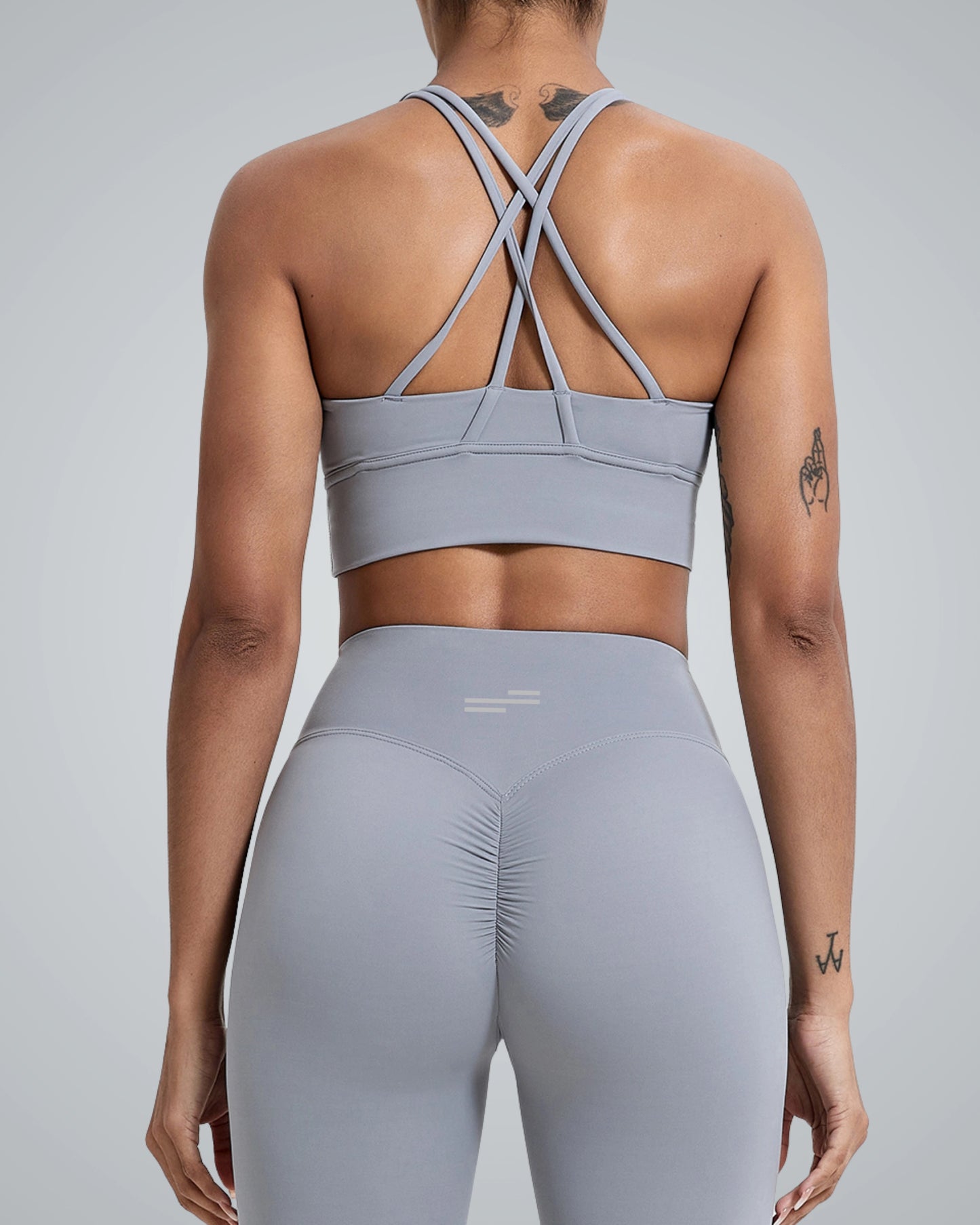 A woman wearing a Bandsam gray sports bra with crisscross straps and matching high-waisted leggings, viewed from the back against a neutral gray background.