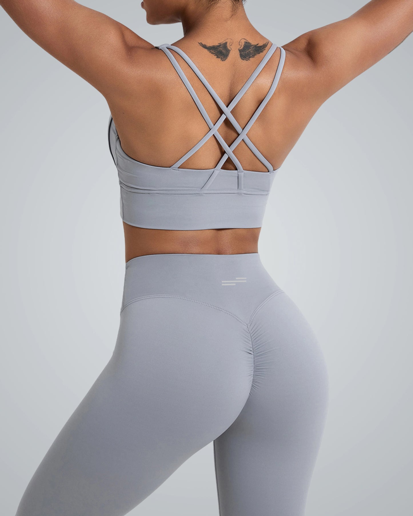 A woman from the back, wearing a Bandsam light gray sports bra with crossed straps and matching high-waisted leggings. She has a tattoo on her upper back, and her athletic figure is highlighted against a light gray background.