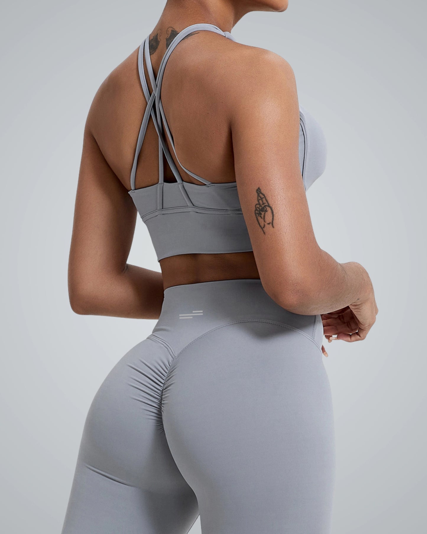 A woman in a gray Bandsam sports bra and leggings, showcasing a back view. The outfit features a stylish crisscross design on the straps and form-fitting fabric that highlights her silhouette. Her tattoo is visible on her upper arm.