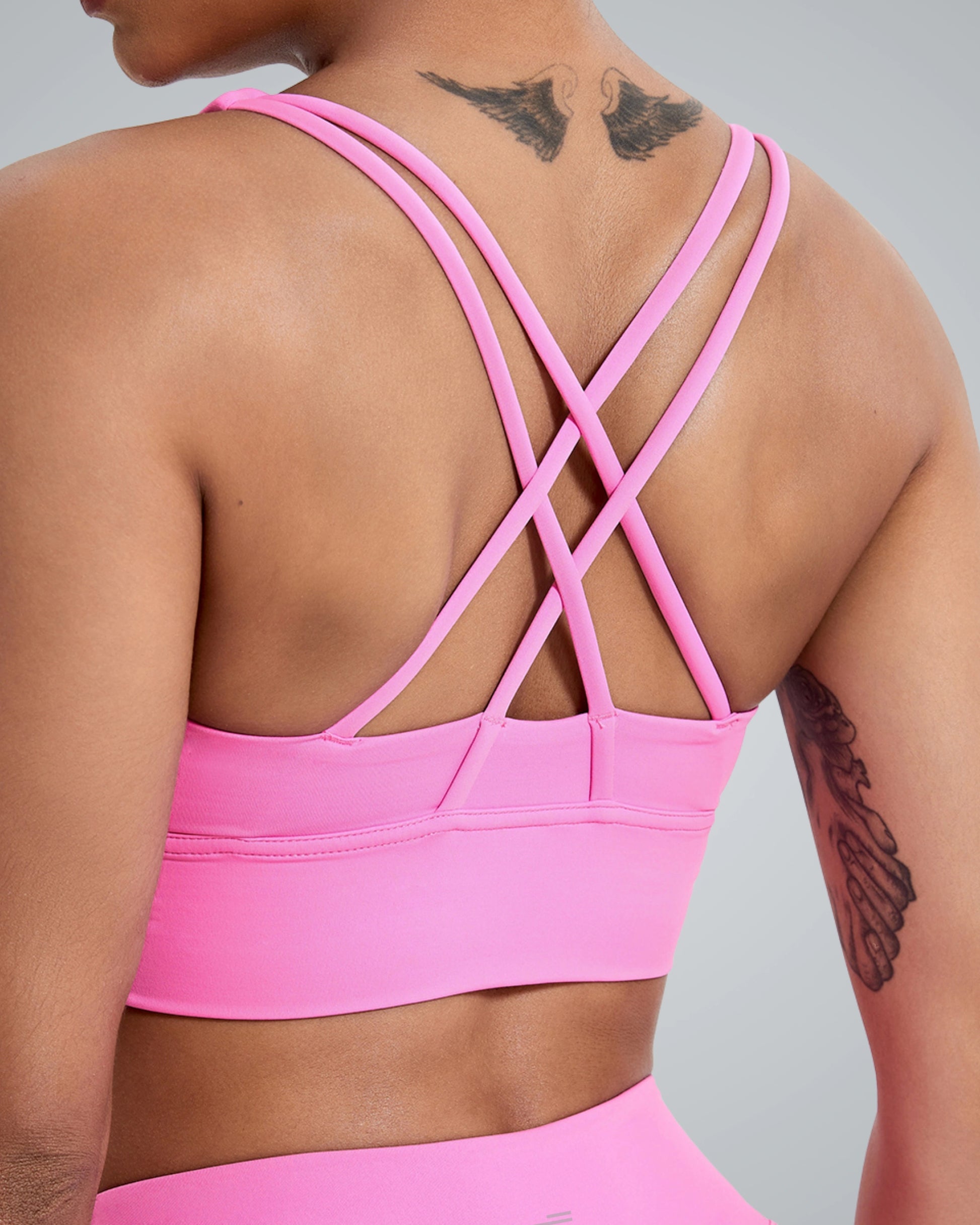 A women wearing a bright Bandsam pink FlexFit Bra with a stylish crisscross design on the back. Visible tattoos are on their upper back and arm, while the neutral gray background highlights this breathable workout bra.