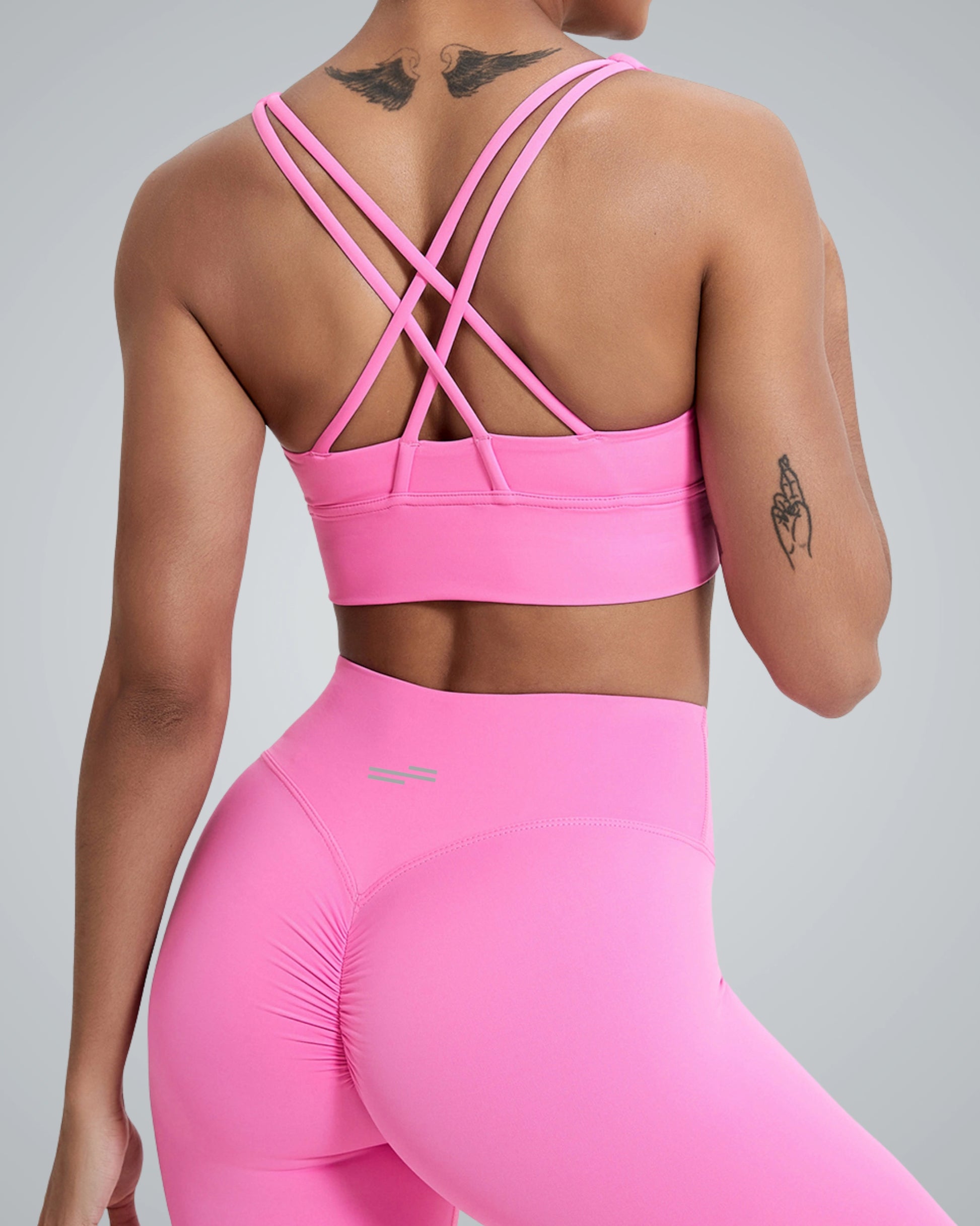 A woman in a Bandsam pink sports bra with crisscross straps and matching high-waisted leggings, showcasing a sporty look from the back. The background is a subtle gray, emphasizing the vibrant color of her outfit.