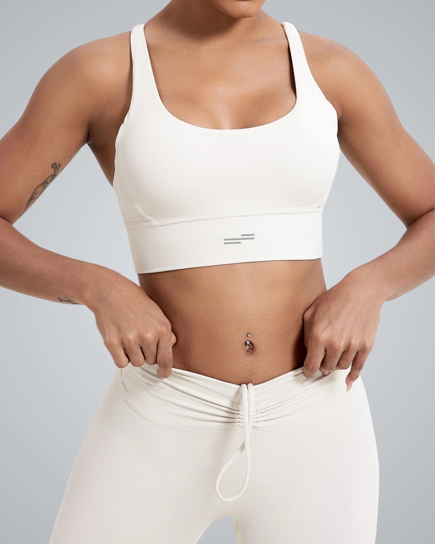 A woman wearing a breathable Bandsam white workout bra and matching high-waisted leggings adjusts the waistband of her leggings. Her midriff and a belly button piercing are visible against a plain light gray background.