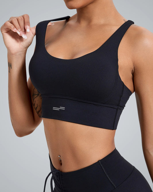 A close-up of a woman wearing a Bandsam sleek black sports bra with wide straps, showcasing a minimalistic design. The backdrop is a smooth gray, highlighting the contours of the garment and the woman's tattooed arm.