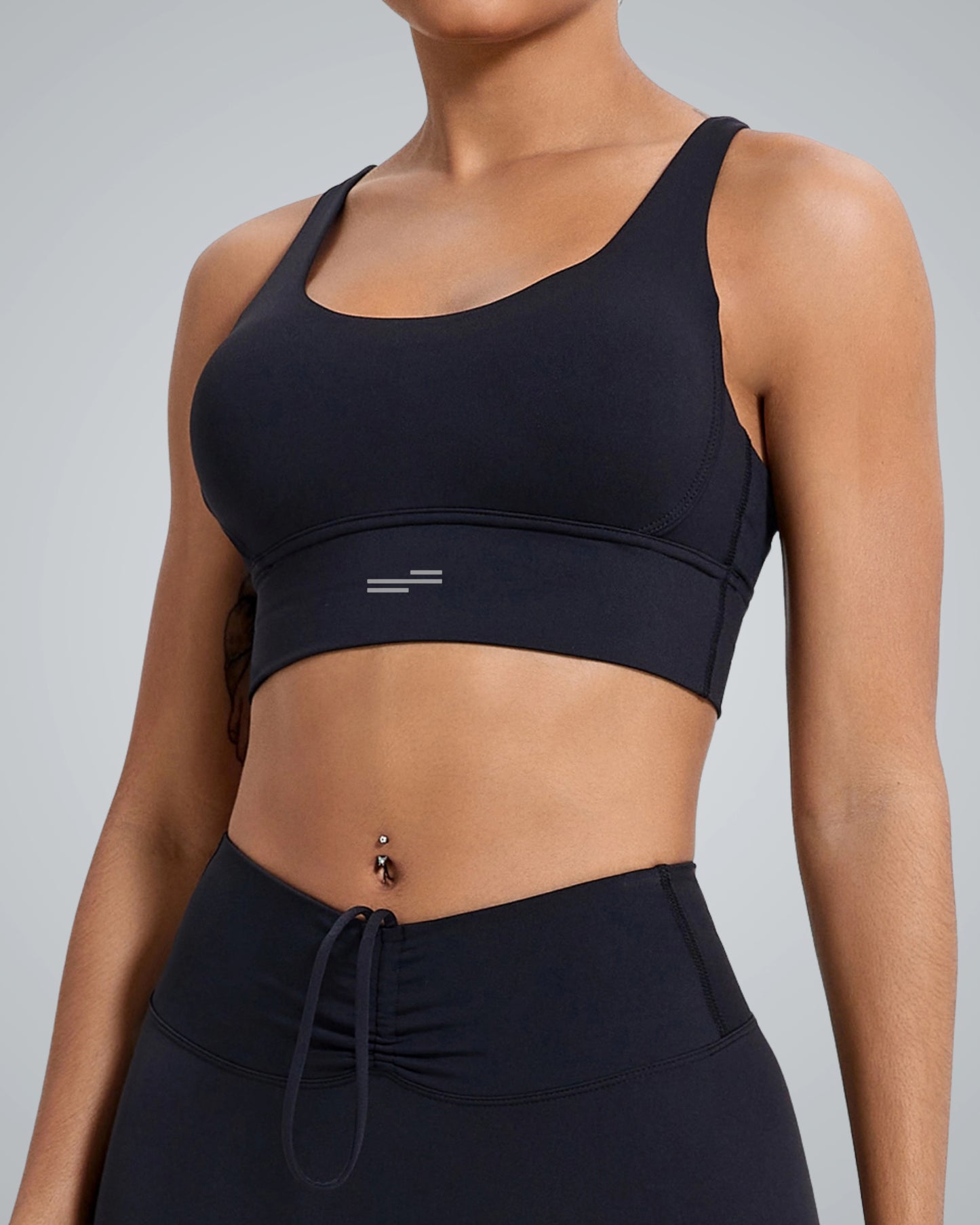 A close-up of a woman's upper body wearing a Bandsam black sports bra and matching high-waisted leggings. The design features a subtle logo on the sports bra, and the model has a piercing on her navel.