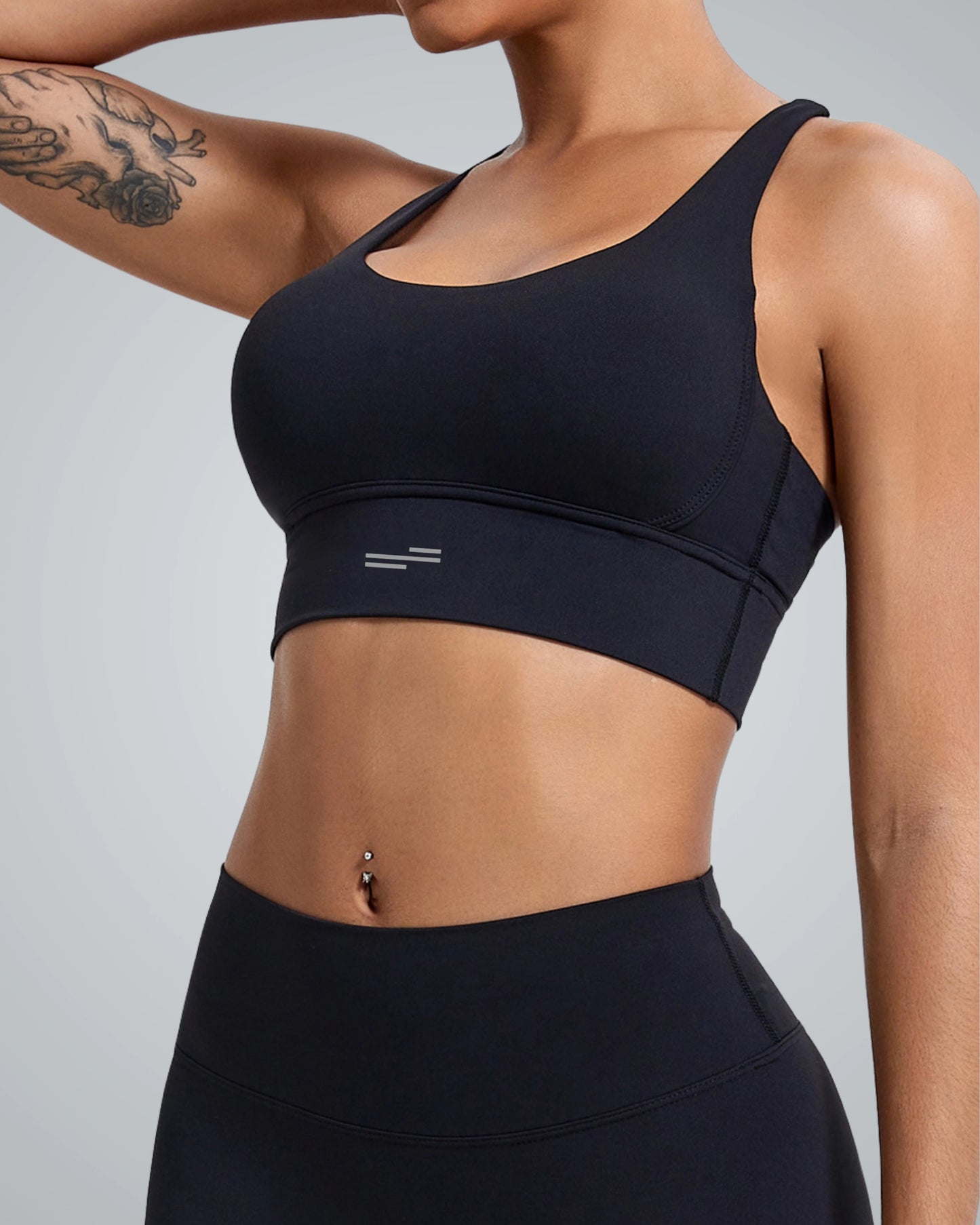 A woman wearing a Bandsam black sports bra, showcasing a fit physique. The bra has a sleek design with a small logo on the front, complemented by matching high-waisted leggings. Her arm features a floral tattoo, and the background is a soft gray, emphasiz