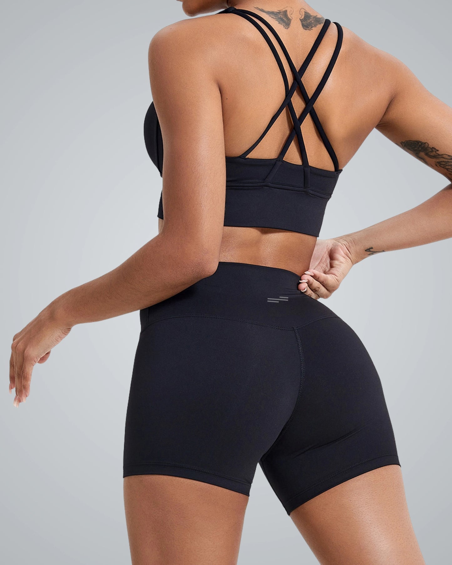 A close-up view of the back of a woman wearing a Bandsam black sports bra with crisscross straps and black high-waisted shorts, showcasing a modern activewear design against a neutral grey background.