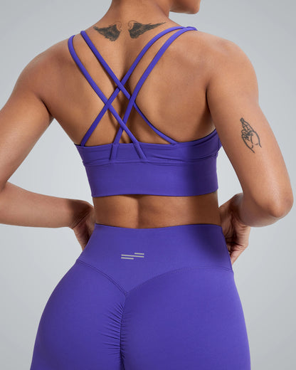 A women wearing a Bandsam purple FlexFit Bra with a crisscross design and matching high-waisted leggings, showing tattoos on their upper back and right arm, stands against a plain light background.