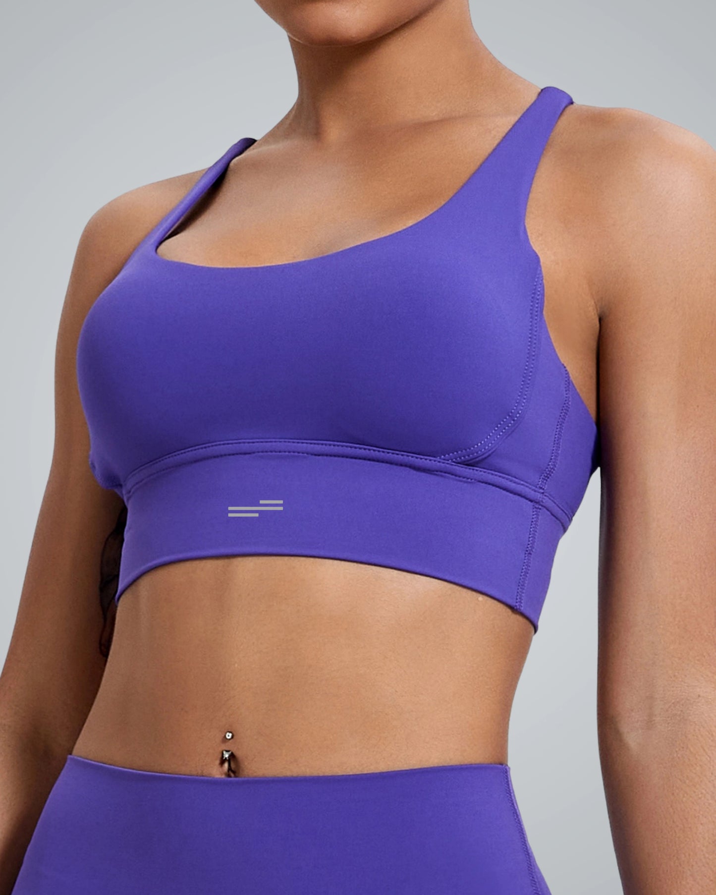 A women wearing a fitted Bandsam purple FlexFit Bra and matching leggings, showing their midriff with a belly button piercing, against a plain light gray background.