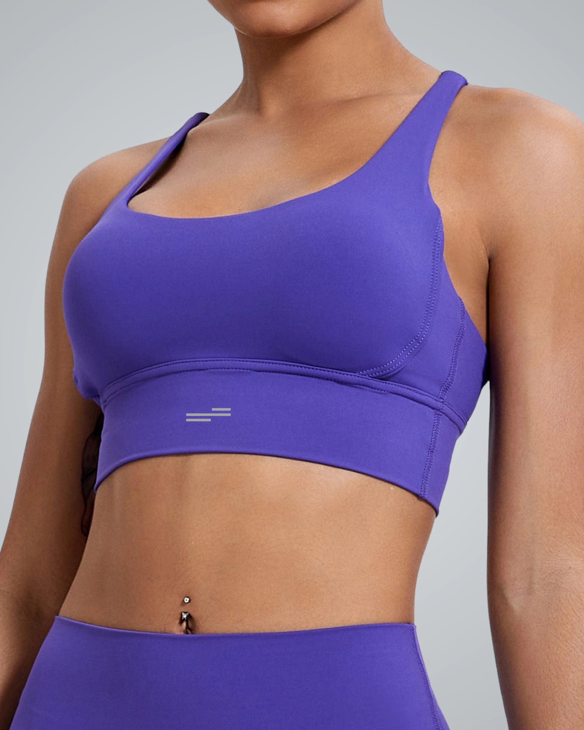 A women wearing a fitted Bandsam purple FlexFit Bra and matching leggings, showing their midriff with a belly button piercing, against a plain light gray background.