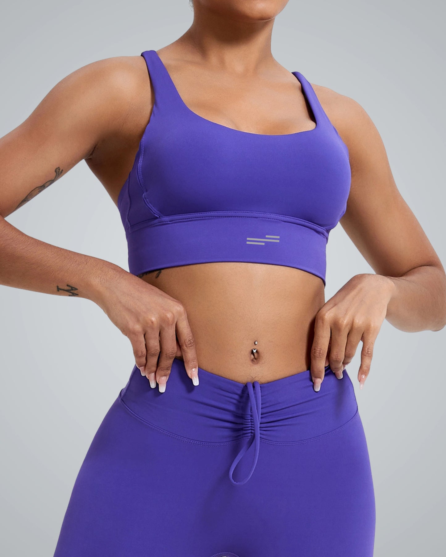 A women wearing a breathable Bandsam workout bra and matching high-waisted leggings stands against a plain background, hands adjusting the waistband. The crisscross design of the bra offers stylish support for yoga. The face is not visible.