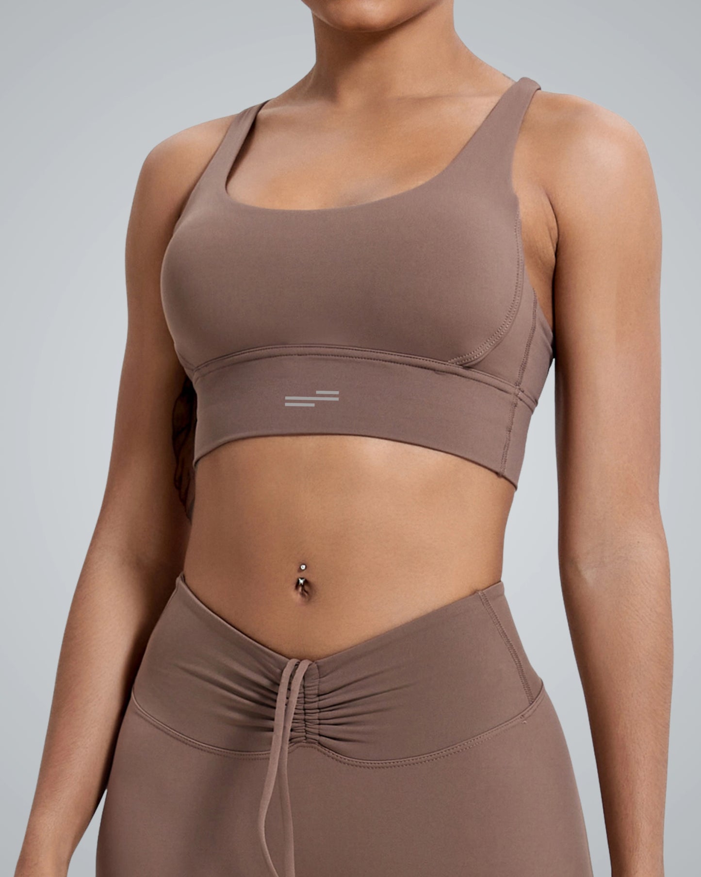 A woman wearing a Bandsam brown sports bra and matching high-waisted leggings, showcasing a minimalist design. The sports bra features a subtle stripe detail, and she is standing in a neutral-toned setting with a smooth background.