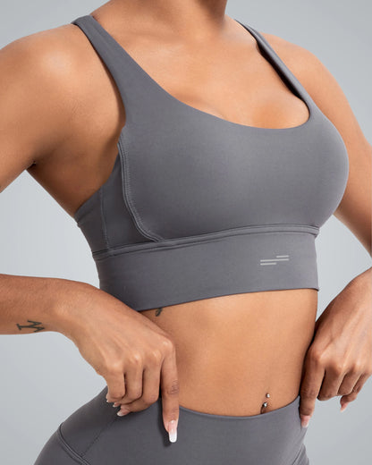 A close-up image of a woman wearing a Bandsam gray sports bra, showcasing its comfortable design with wide straps. She has her hands positioned at her waist, emphasizing the fit of the garment against a smooth gray background.