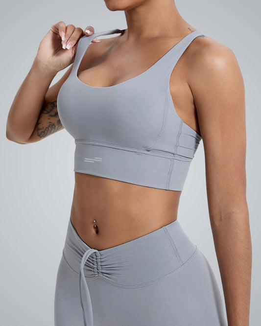 A woman adjusting a light gray Bandsam sports bra with a subtle logo detail, paired with matching high-waisted leggings featuring a drawstring design, showcasing a sleek and supportive activewear set.