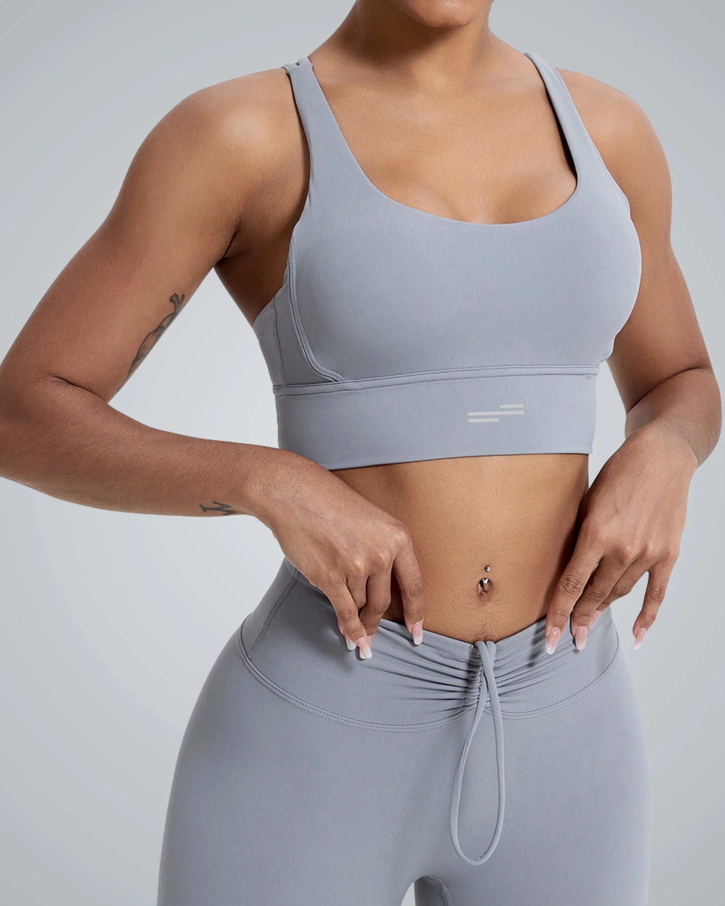 A women wearing a Bandsam light gray sports bra and matching high-waisted leggings, posing with hands on their hips, showcasing a toned midriff. The background has a soft gray gradient, enhancing the athletic wear.
