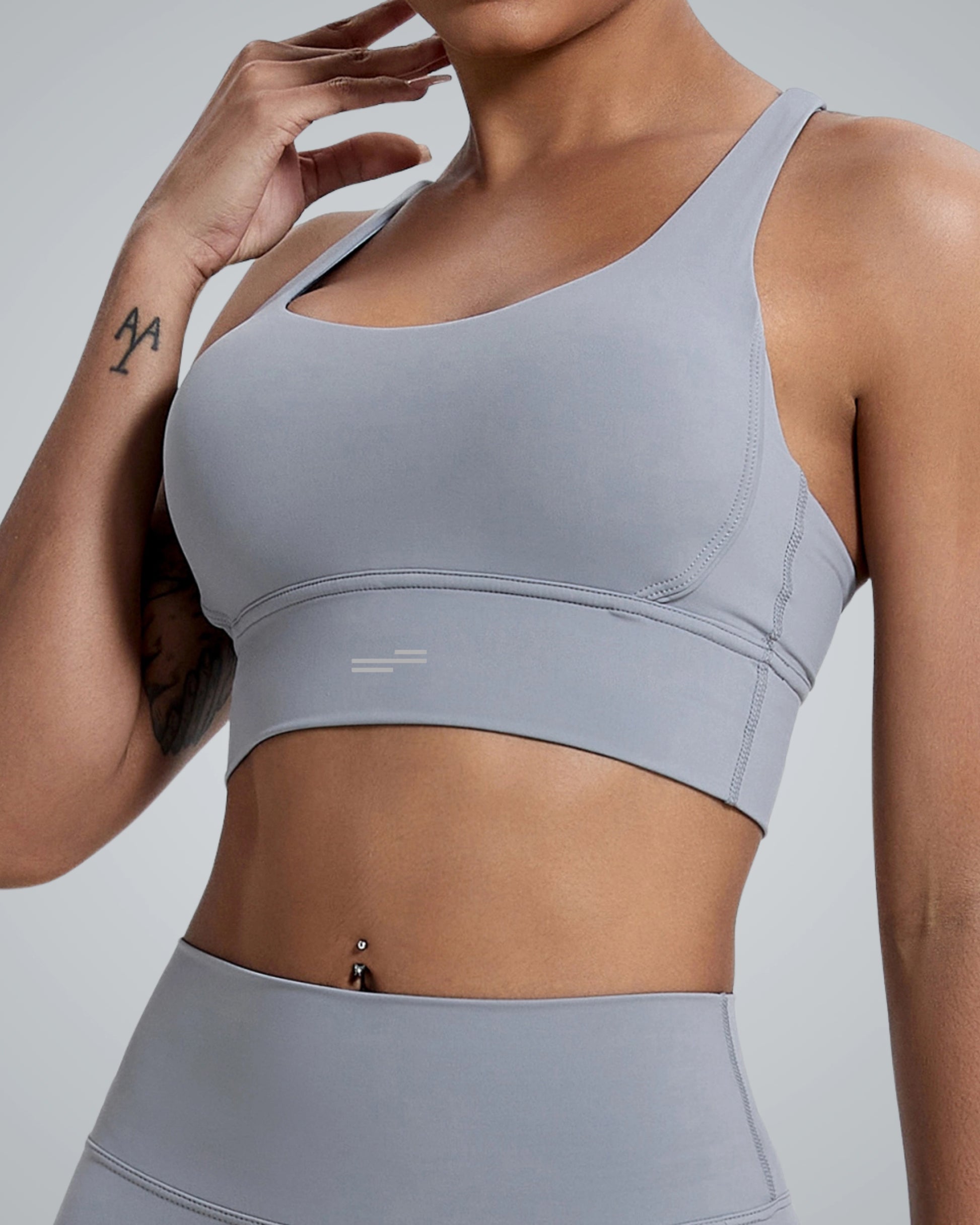 A close-up of a woman wearing a Bandsam light gray sports bra, showcasing a sleek design with a round neckline and racerback style. The background is neutral, emphasizing the activewear and the woman's toned physique.