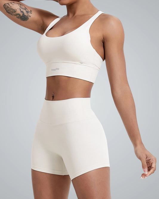 A woman models a Bandsam white FlexFit Bra and matching high-waisted shorts against a plain grey background. The breathable workout bra features a subtle crisscross design. She has a tattoo on her upper arm and a navel piercing. Her face is not visible.