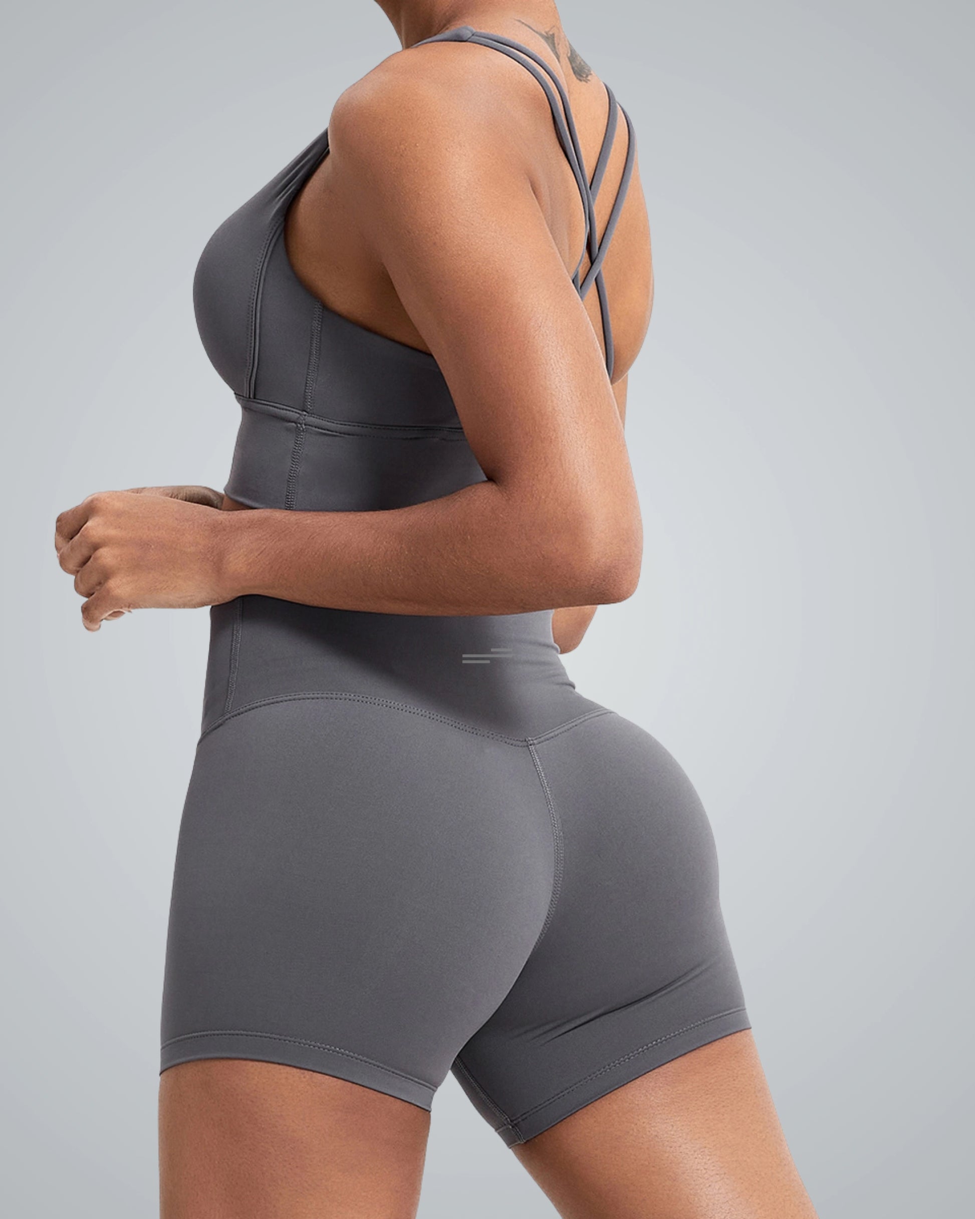 A woman wearing a Bandsam gray athletic bra with short with crisscross straps, posing sideways. The outfit highlights her silhouette against a smooth gray background, emphasizing fitness and style.