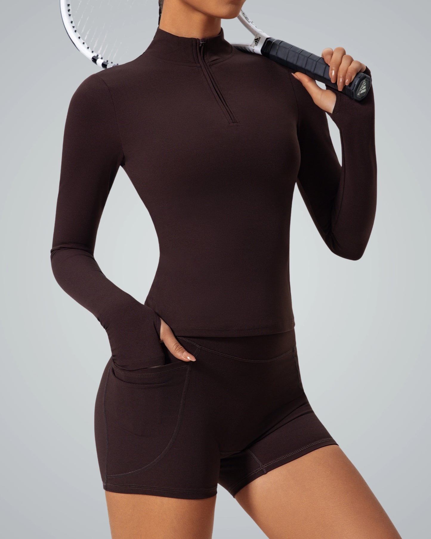 Woman in brown seamless gym set holding tennis racket, featuring long sleeve zip-up and shorts, part of Bandsam's FluidForce Collection.