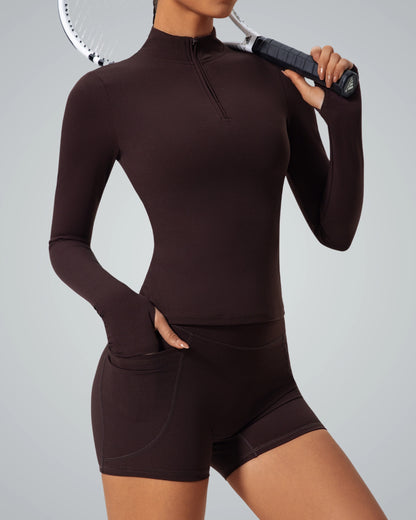 Woman in brown seamless gym set holding tennis racket, featuring long sleeve zip-up and shorts, part of Bandsam's FluidForce Collection.
