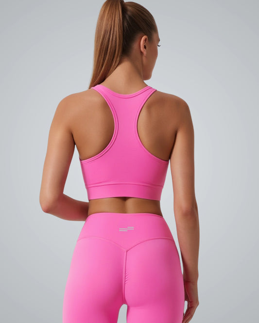 A woman with long straight brown hair in a ponytail is shown from the back, wearing a Bandsam bright pink GlowMotion Bra and matching high-waisted leggings against a plain, light gray background.