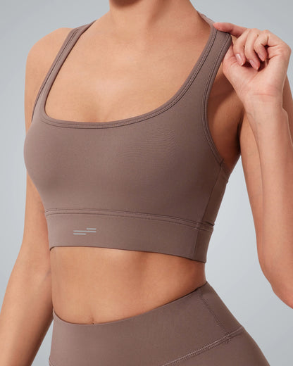 A woman wearing a Bandsam GlowMotion Bra and matching high-waisted leggings poses against a plain gray background, holding one strap of her high impact sports bra with her right hand. Her face is not visible.