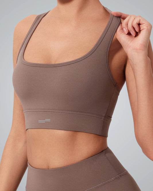 A woman wearing a Bandsam GlowMotion Bra and matching high-waisted leggings poses against a plain gray background, holding one strap of her high impact sports bra with her right hand. Her face is not visible.