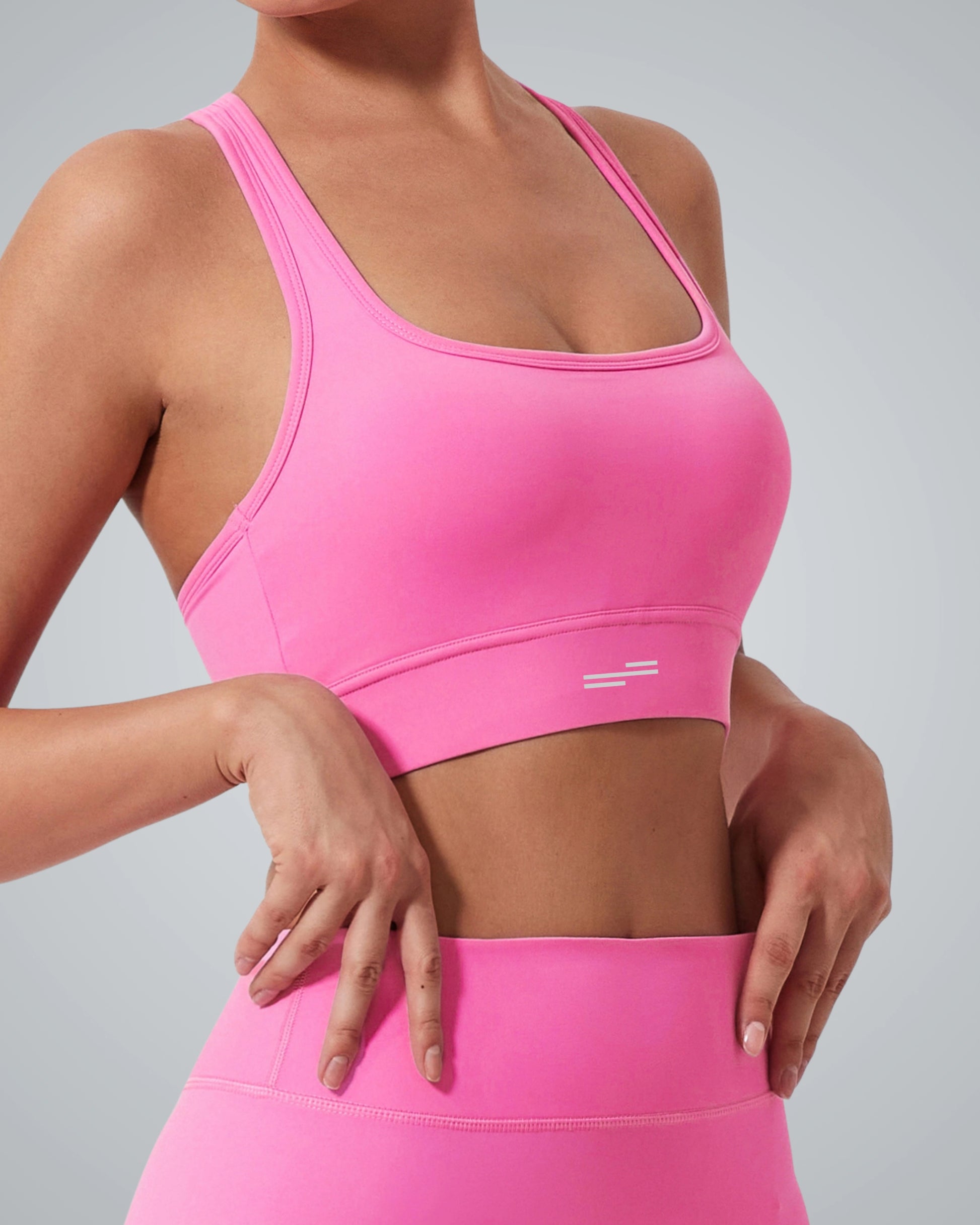 A woman wearing a Bandsam bright pink GlowMotion Bra, known for its support as a high impact sports bra, and matching high-waisted leggings stands against a plain gray background, with one hand resting on her hip and the other slightly raised.