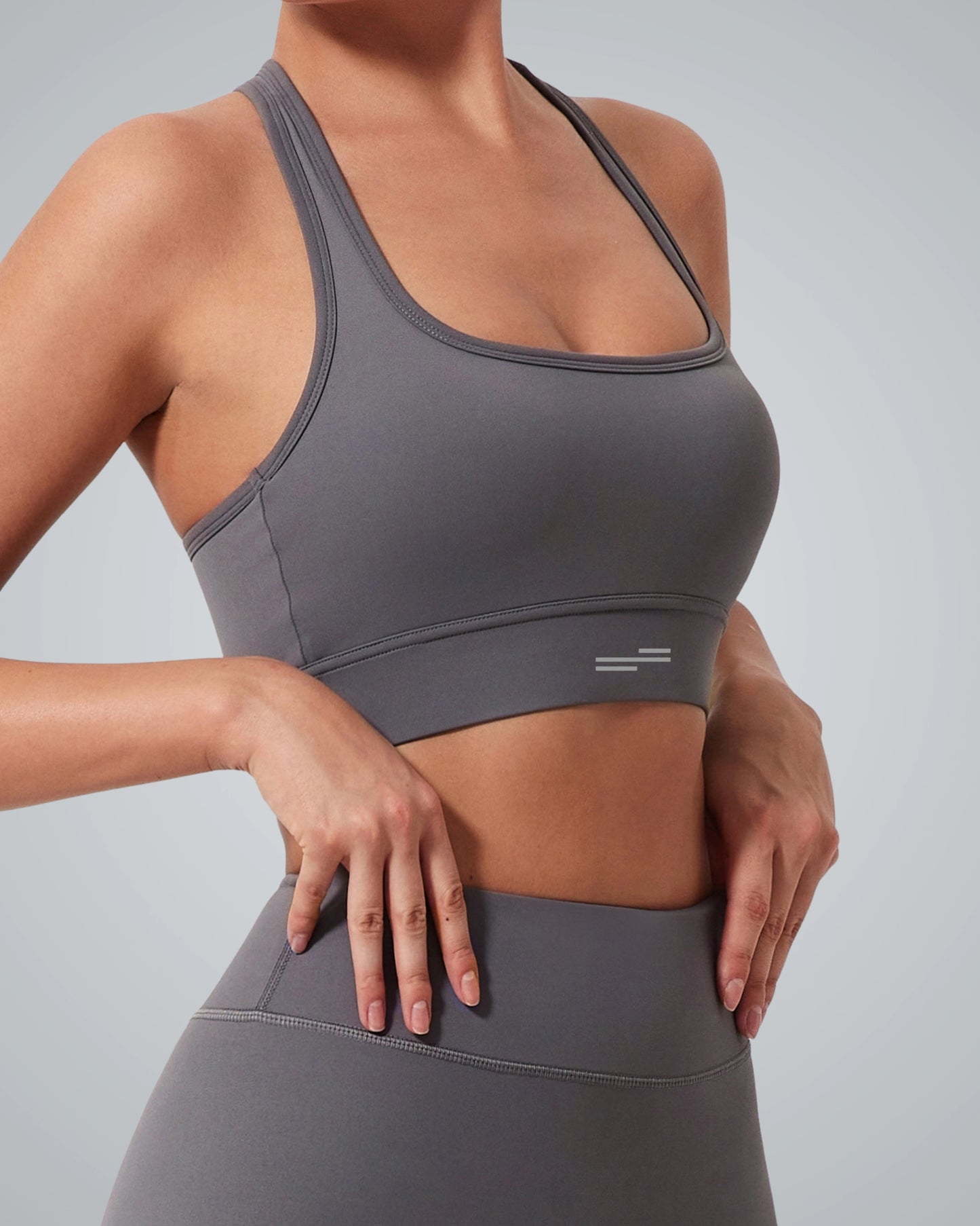 A woman wearing a Bandsam gray high impact sports bra and matching high-waisted leggings stands with her hands resting on her hips, ready to take on any active routine against a plain background.