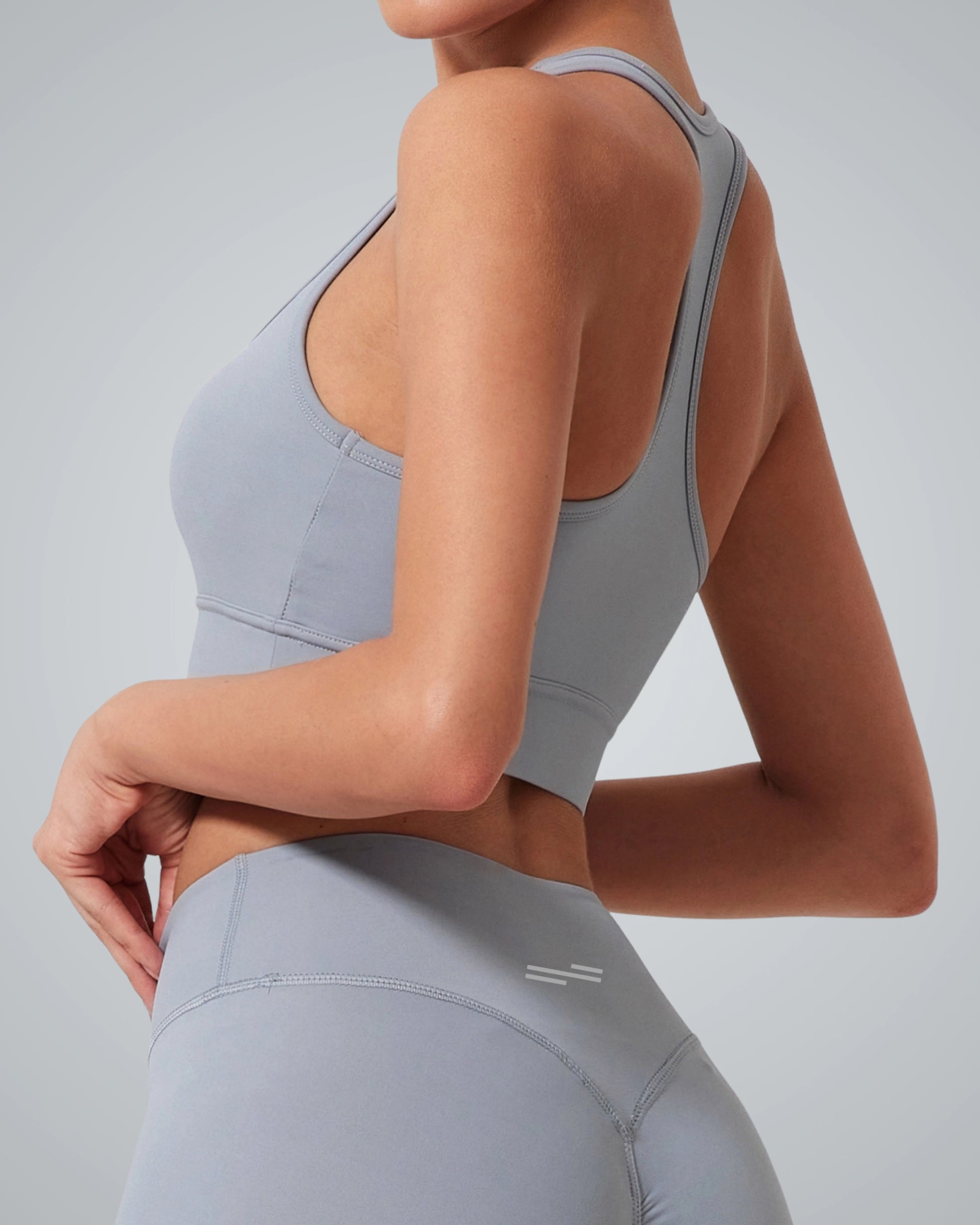 A woman wearing a Bandsam GlowMotion Bra—a light gray, high impact sports bra—and matching high-waisted leggings stands with one hand on her hip, showing the back and side of her supportive athletic outfit against a plain background.