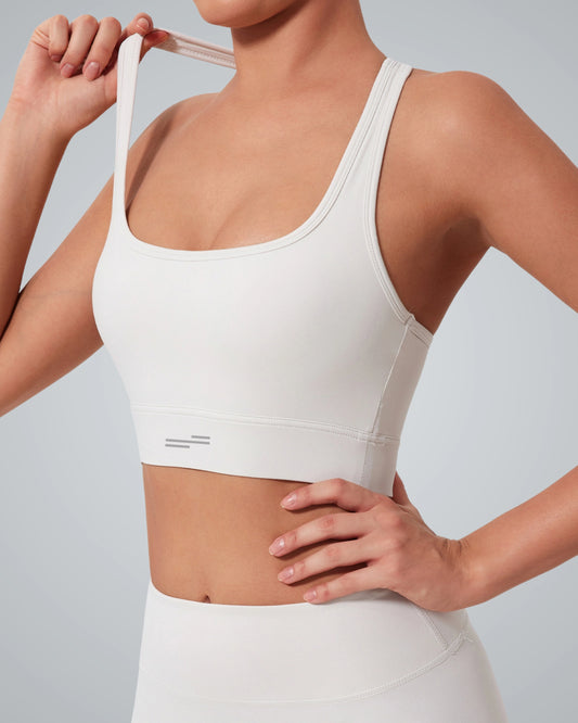 A woman wearing a light beige high impact sports bra and matching high-waisted leggings stands with one hand on her hip and the other holding a shoulder strap, showcasing the support of the Bandsam GlowMotion Bra against a plain gray background.