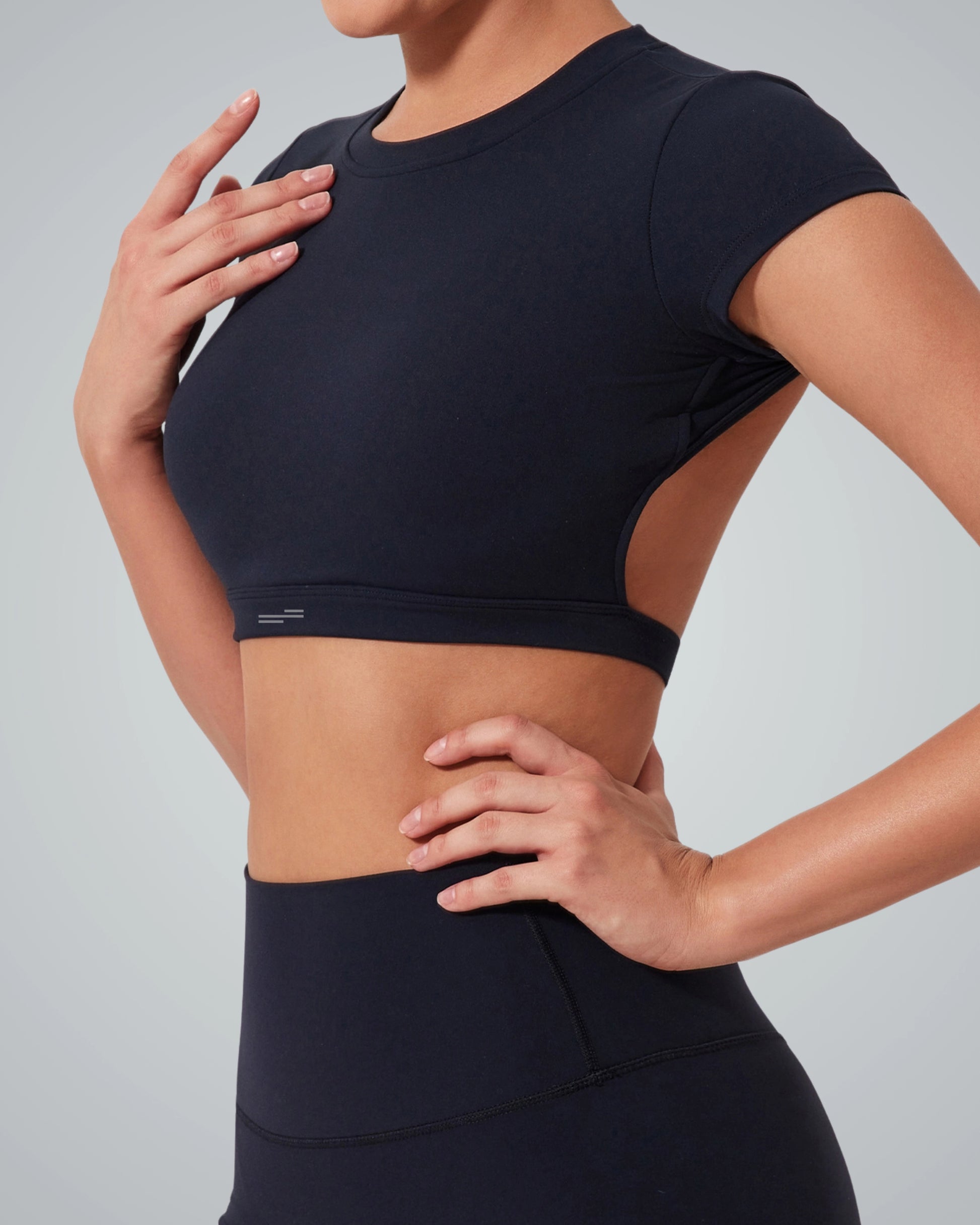 A woman wearing a Bandsam fitted short-sleeve ScarletFlow Top with an open back and high-waisted black leggings, posing with one hand on her hip and the other resting on her upper chest against a plain gray background—perfect for your exercise wardrobe.