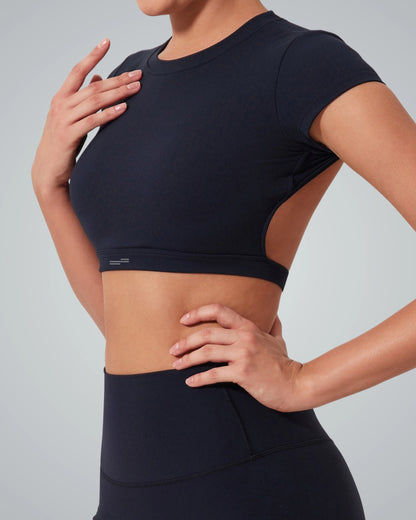 A woman wearing a Bandsam fitted short-sleeve ScarletFlow Top with an open back and high-waisted black leggings, posing with one hand on her hip and the other resting on her upper chest against a plain gray background—perfect for your exercise wardrobe.