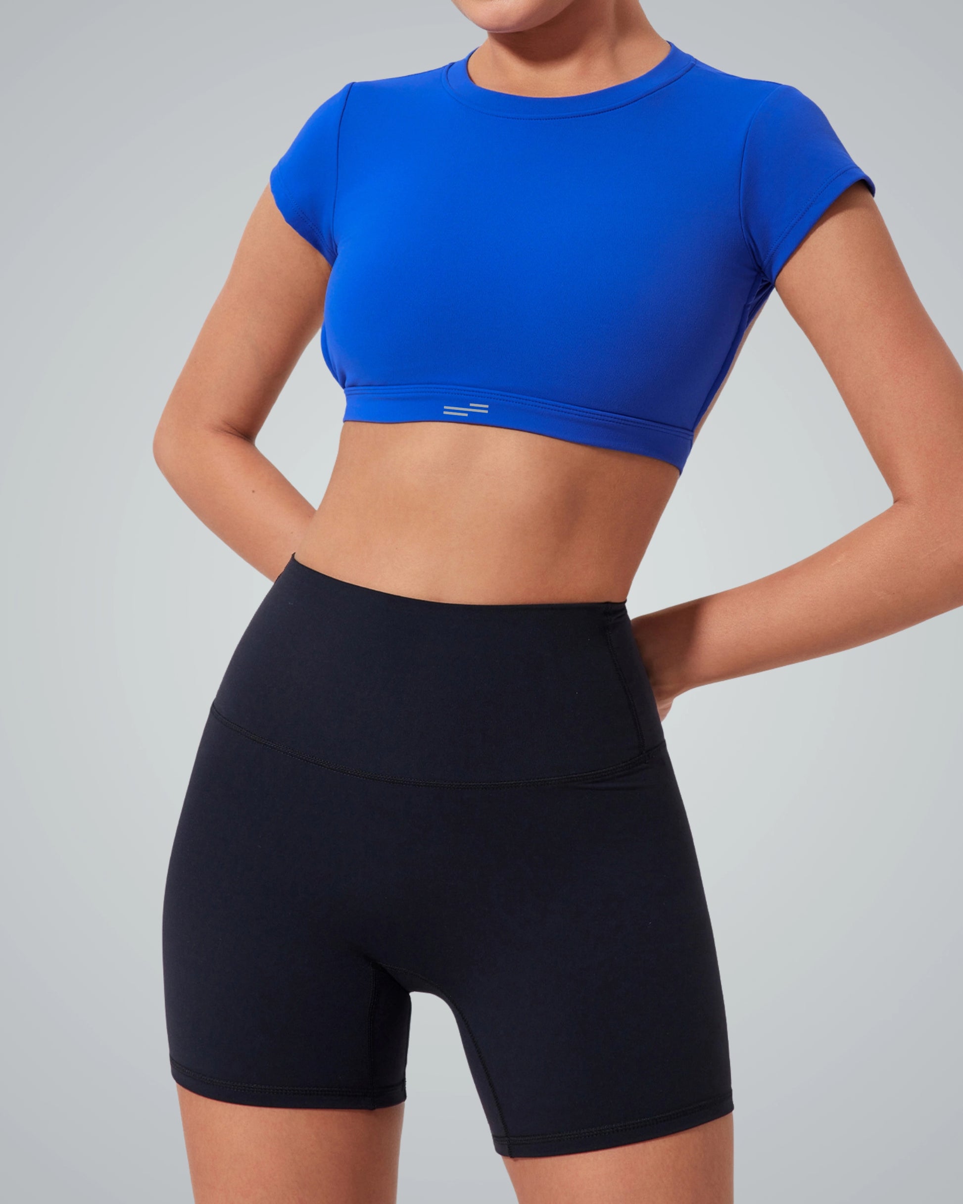 A woman poses in a Bandsam bright blue open back top and high-waisted black biker shorts against a plain light gray background—an ideal look for an active life. Her hands rest on her hips, and her head is out of the frame.