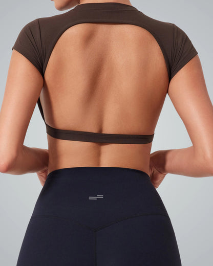 A women is shown from the back wearing a Bandsam brown ScarletFlow Top with an open back and high-waisted black leggings. The top exposes most of the back, making it a chic addition to any exercise wardrobe. The background is plain light gray.