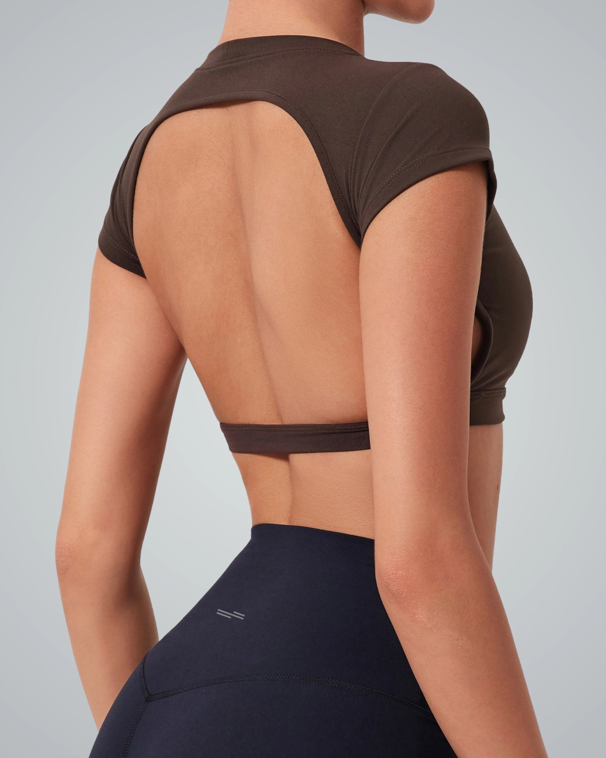 A women is shown from the back wearing a Bandsam brown ScarletFlow Top with an open back and high-waisted black leggings. The top exposes most of the back, making it a chic addition to any exercise wardrobe. The background is plain light gray.