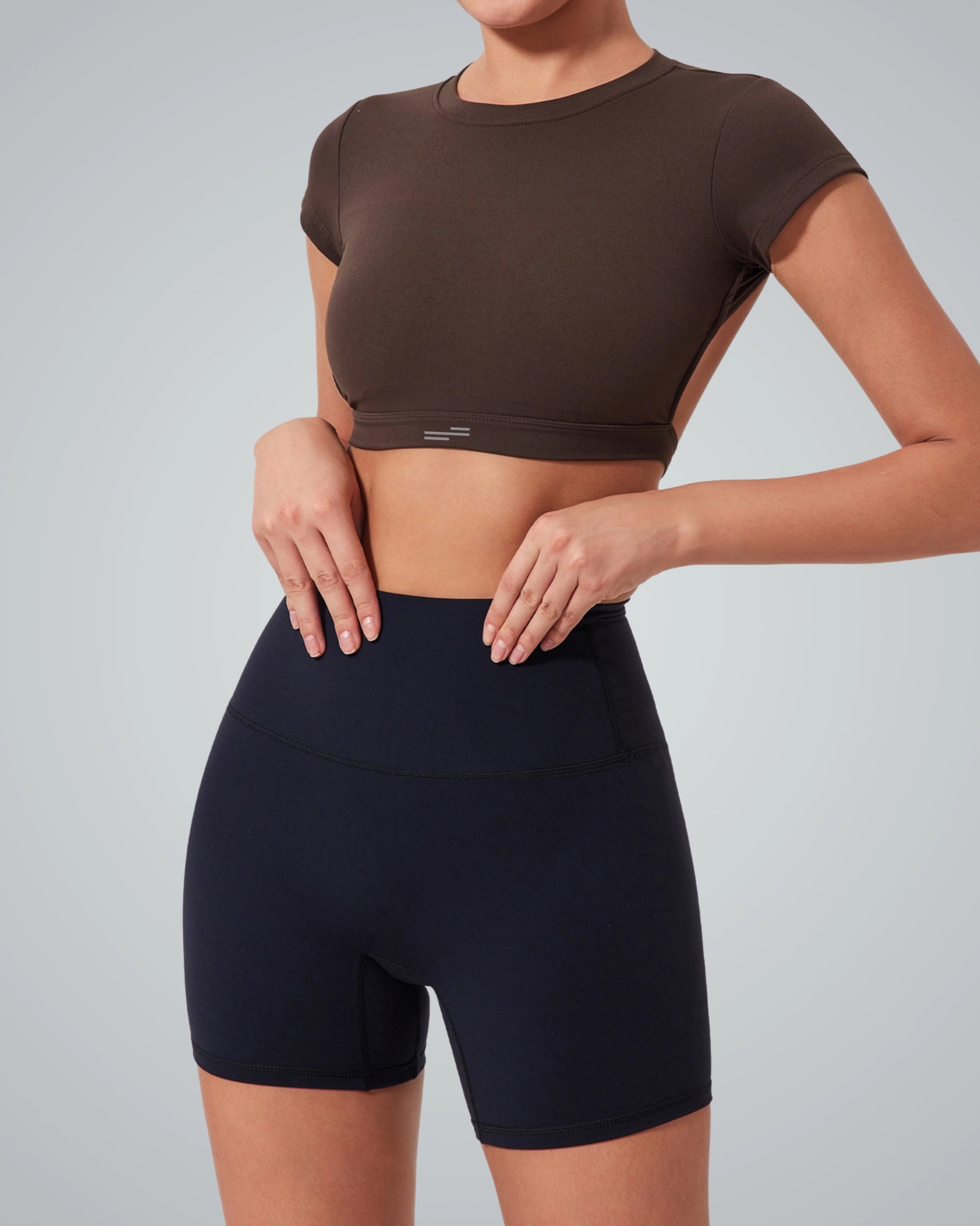 A women wearing a Bandsam short-sleeve, open back brown crop top and high-waisted black biker shorts stands against a plain light gray background. Only the torso and upper legs are visible, perfect for an exercise wardrobe refresh.